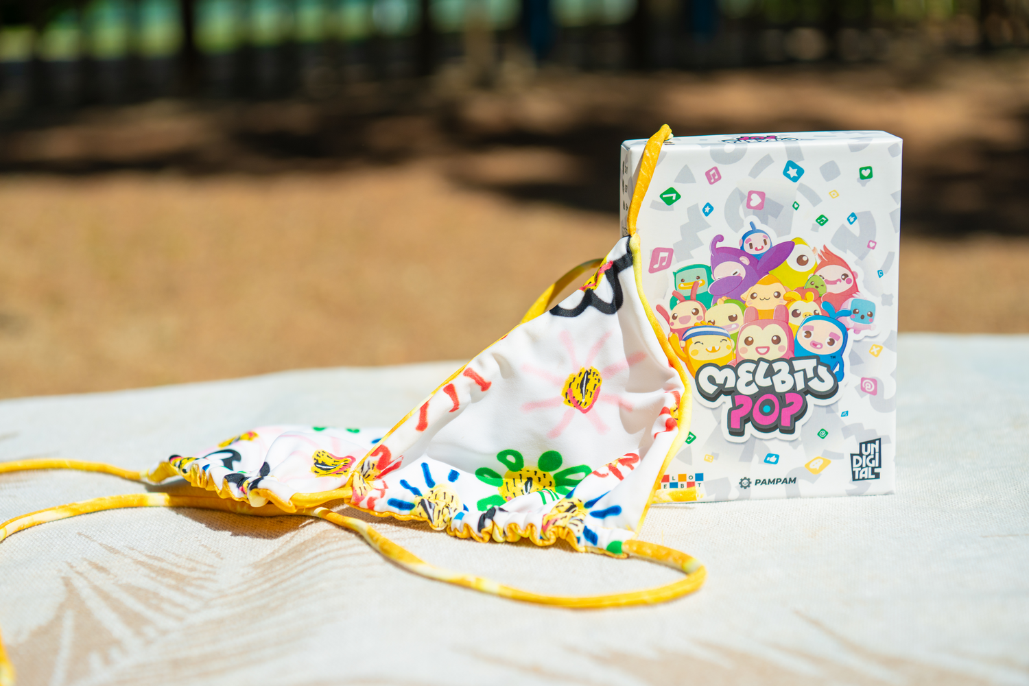 Un bikini colorido y la caja del juego Melbits Pop sobre una manta clara al aire libre.