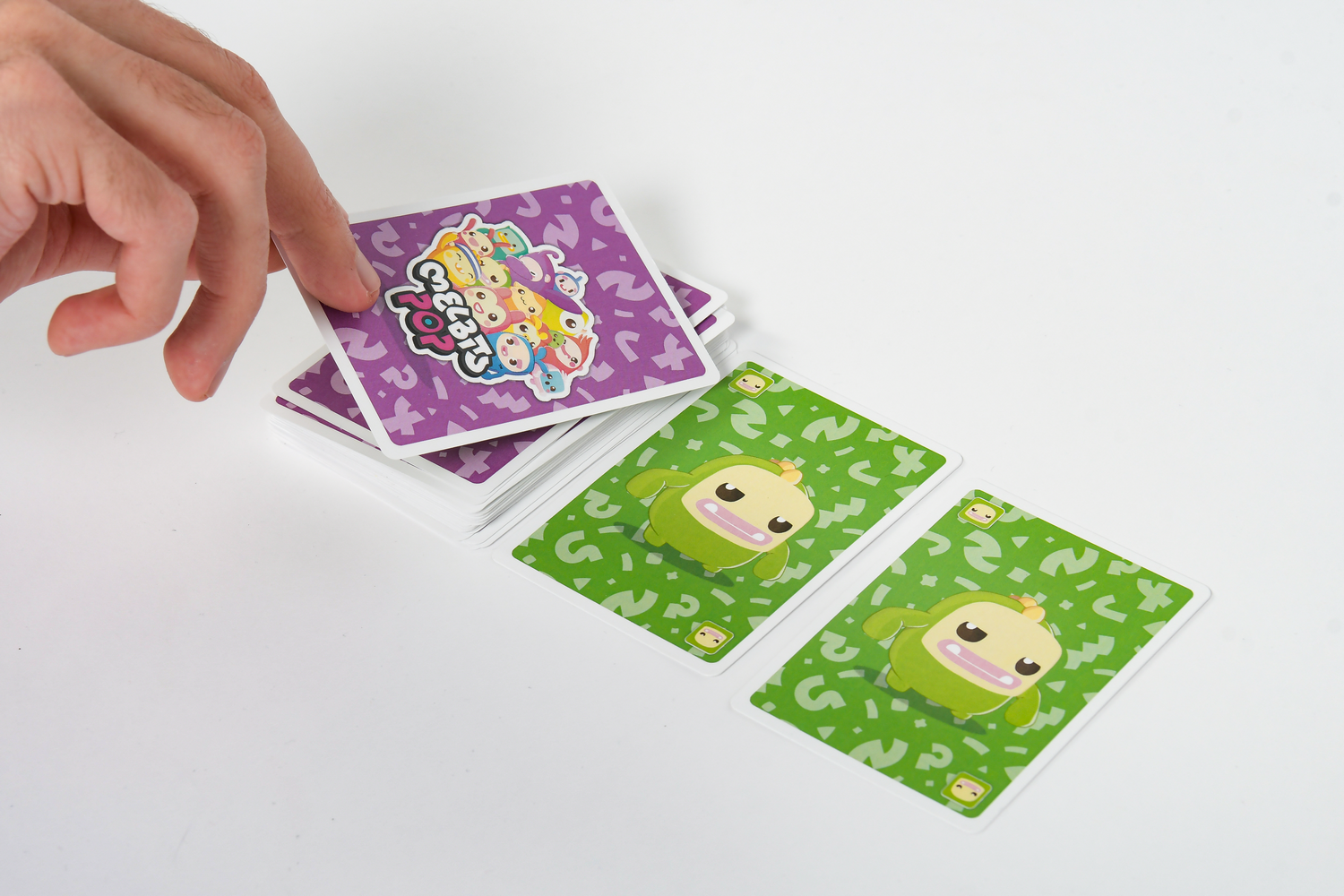 Una mano roba una carta morada de Melbits Pop de un mazo, junto a dos cartas verdes en una mesa blanca.