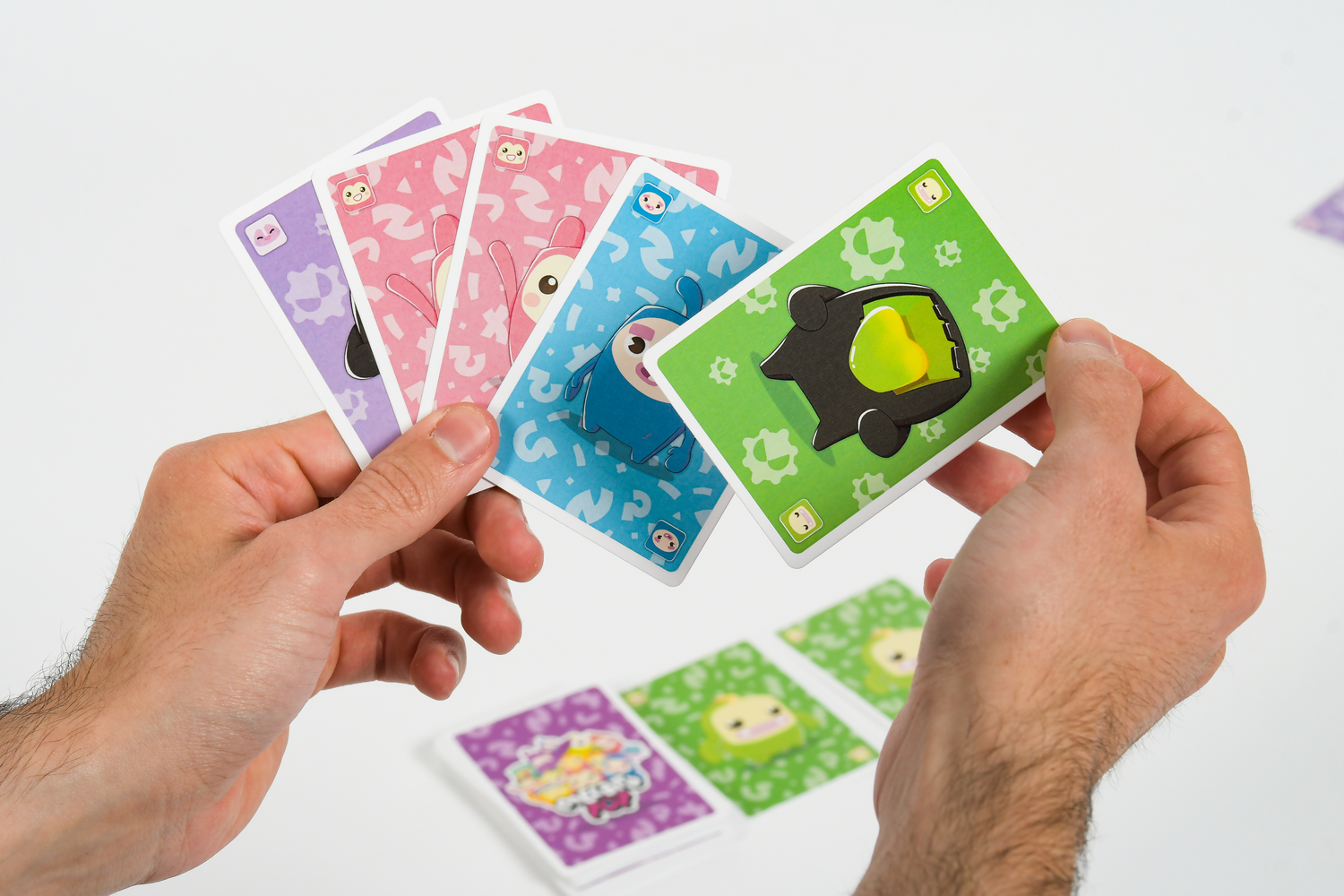 Manos sostienen cartas de juego coloridas con personajes, con más cartas en una mesa blanca.