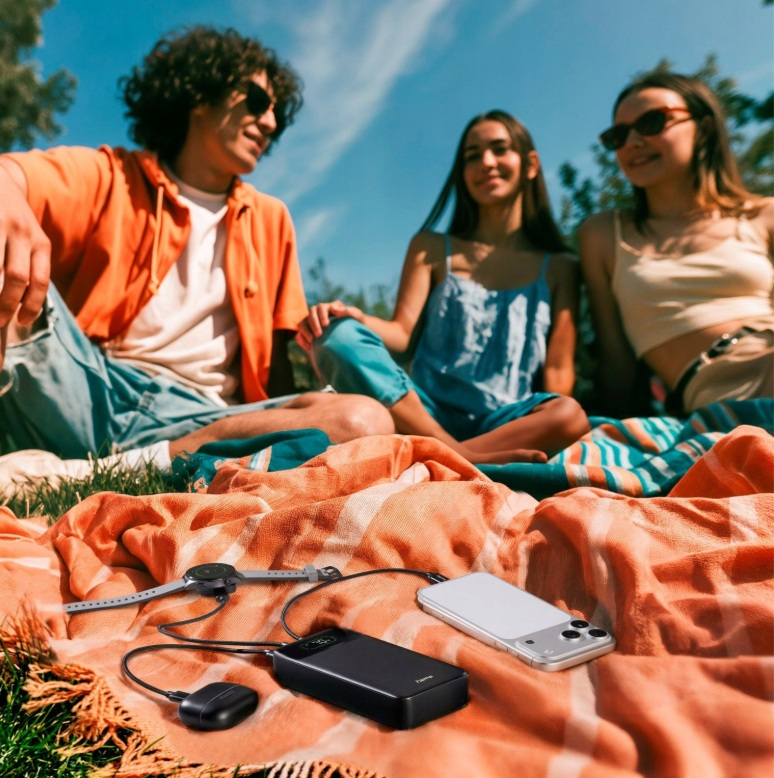 Trzech przyjaciół na kocu na dworze, podczas gdy powerbank ładuje zegarek, słuchawki i telefon.