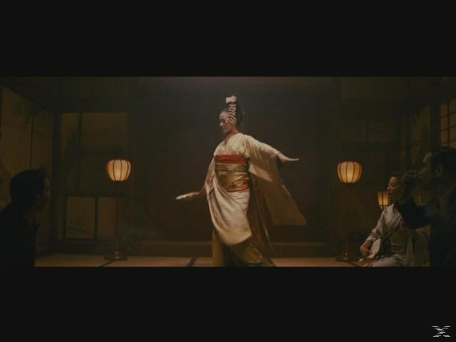 Eine Geisha in einem hellen Kimono tanzt in einem traditionellen Raum mit Lichtern und Zuschauern.