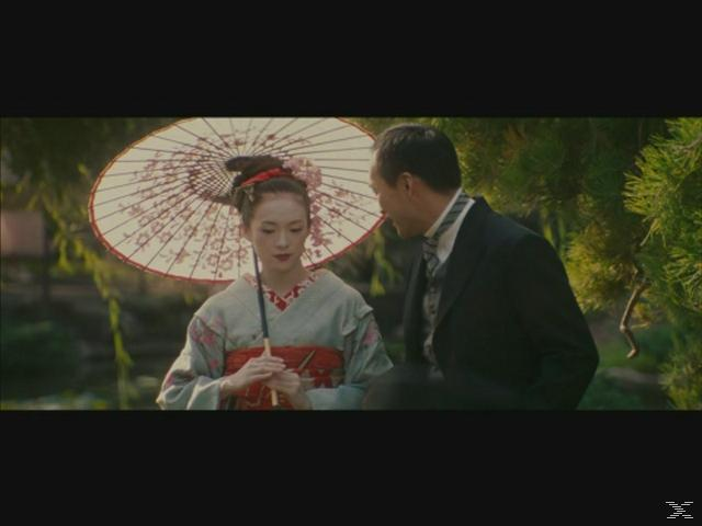 Eine Geisha mit rotem Regenschirm spricht mit einem Mann im Anzug im Freien.