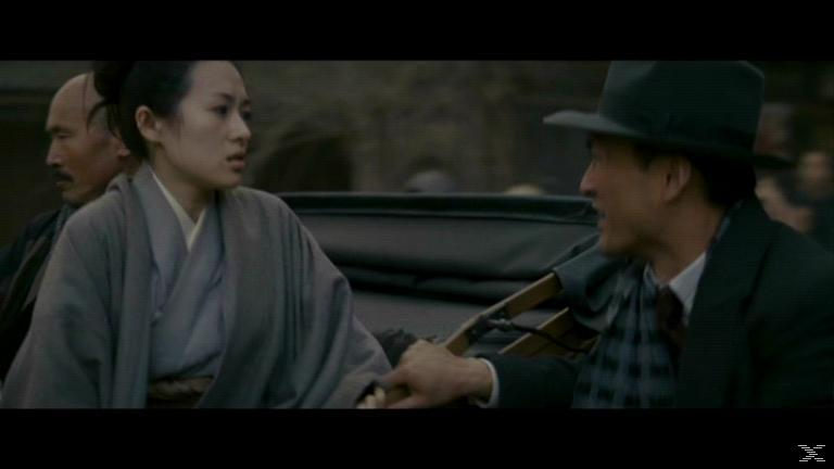 Eine Frau im grauen Kimono sitzt in einer Sänfte und spricht mit einem Mann im Anzug.