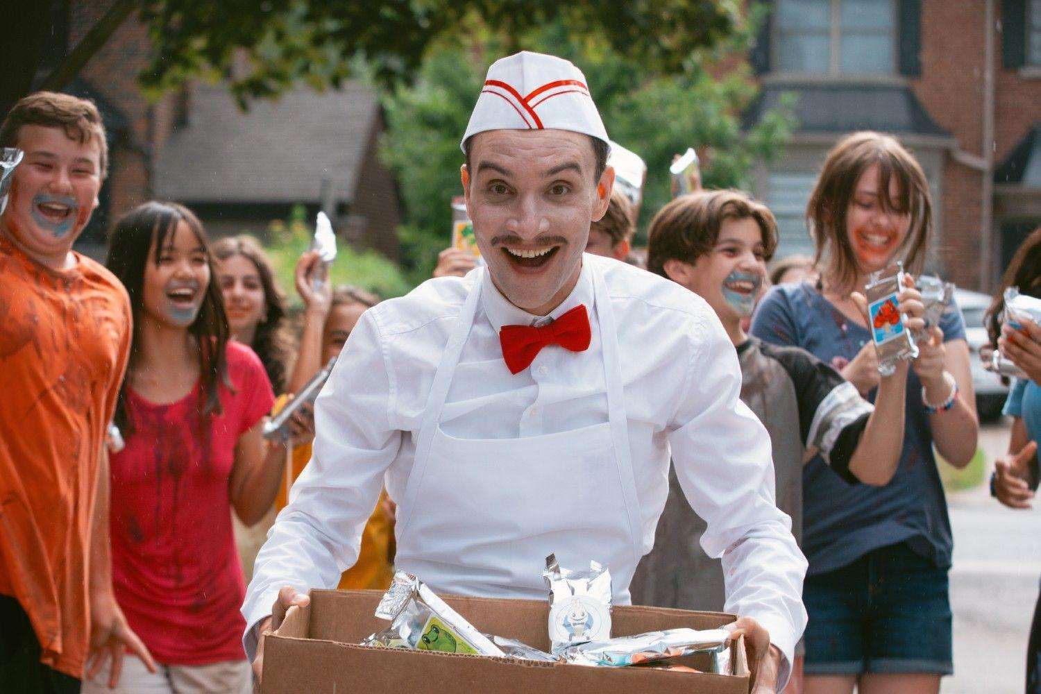 Ein glücklicher Mann in Arbeitskleidung trägt eine Kiste mit Snacks, umgeben von jubelnden Jugendlichen draußen.