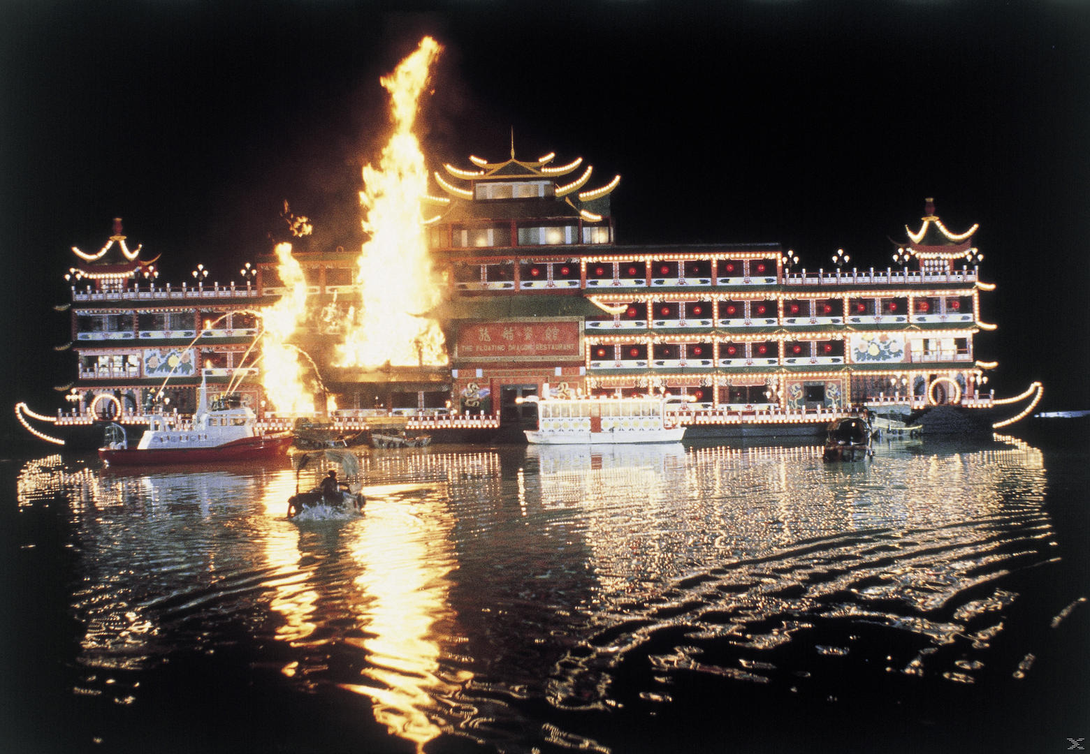 Ein großes, beleuchtetes chinesisches schwimmendes Restaurant steht nachts in Flammen, während Boote im Wasser treiben.