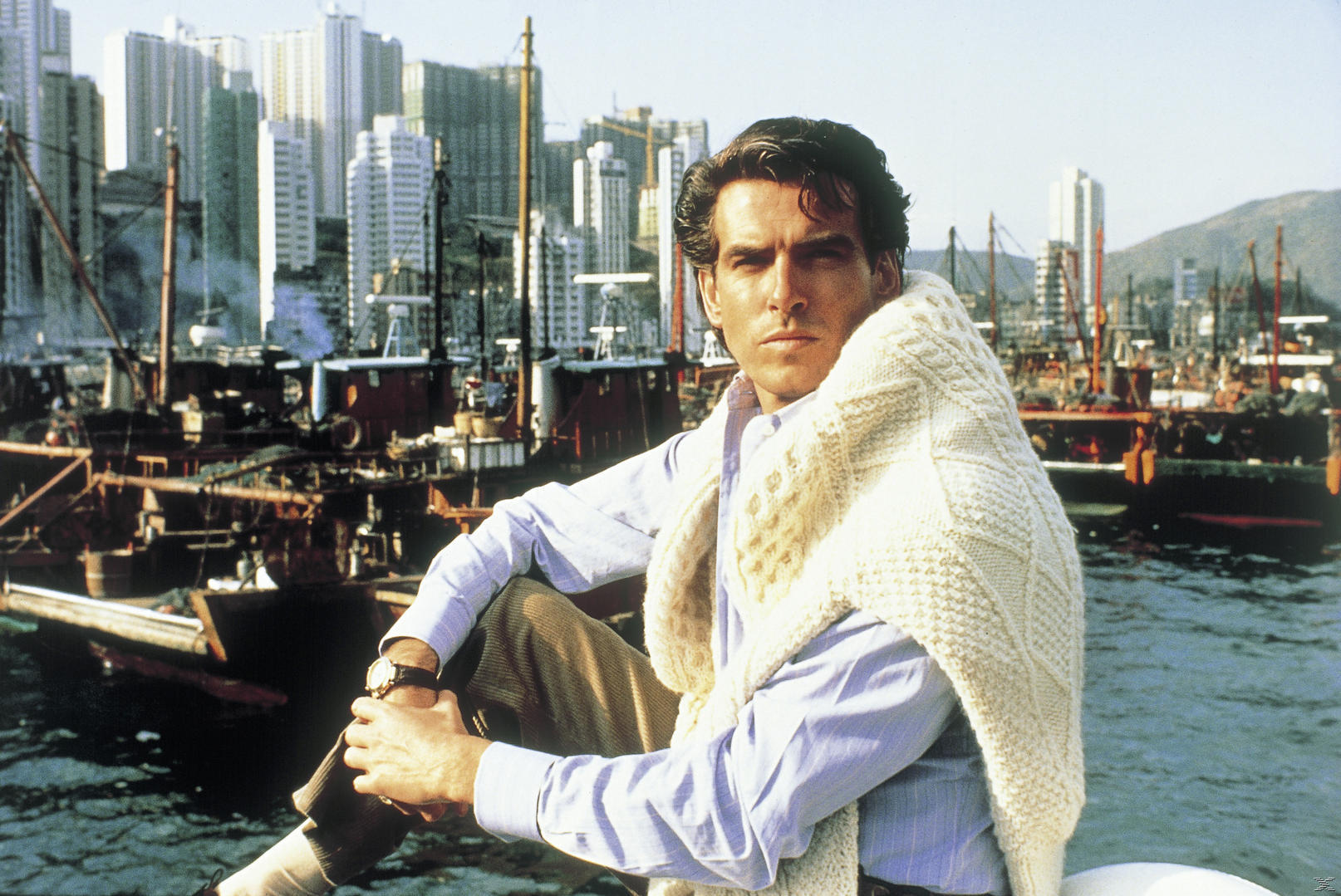 Ein Mann mit weißem Pullover sitzt am Hafen, im Hintergrund sind Schiffe und eine Skyline.