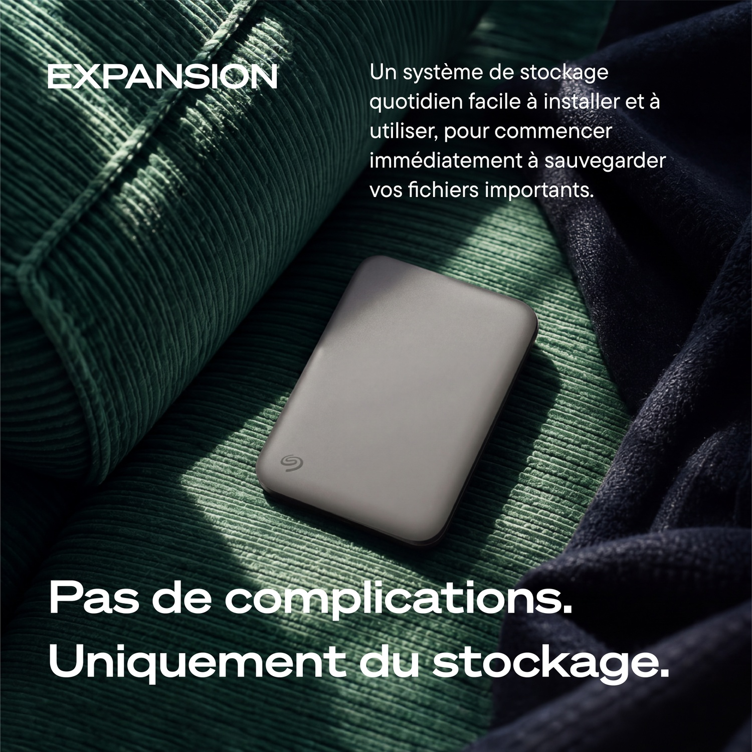Un disque dur externe Seagate gris repose sur un canapé en velours côtelé vert près d'une couverture.
