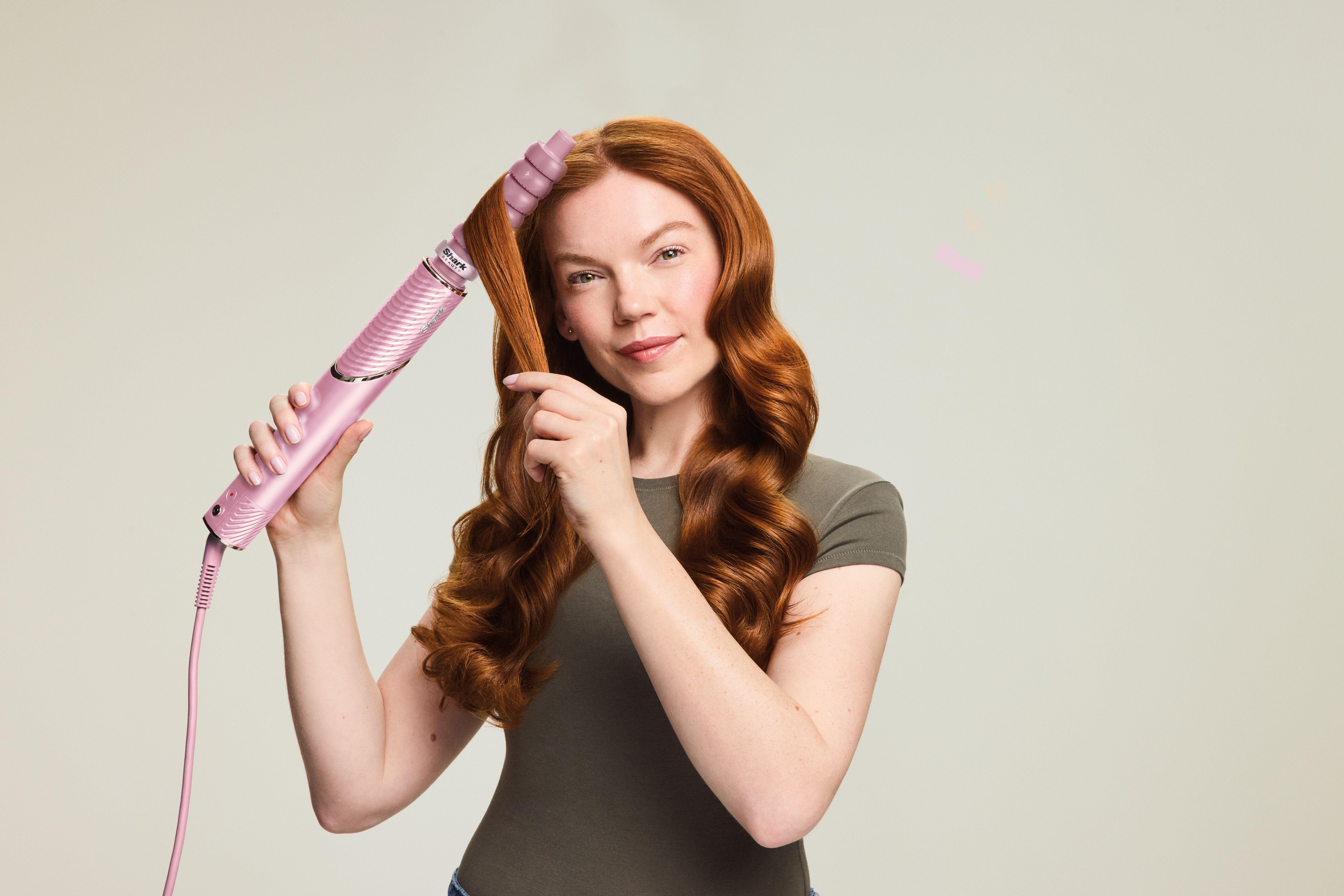 Eine rothaarige Frau lächelt und nutzt einen pinken Lockenstab für ihre Haare.