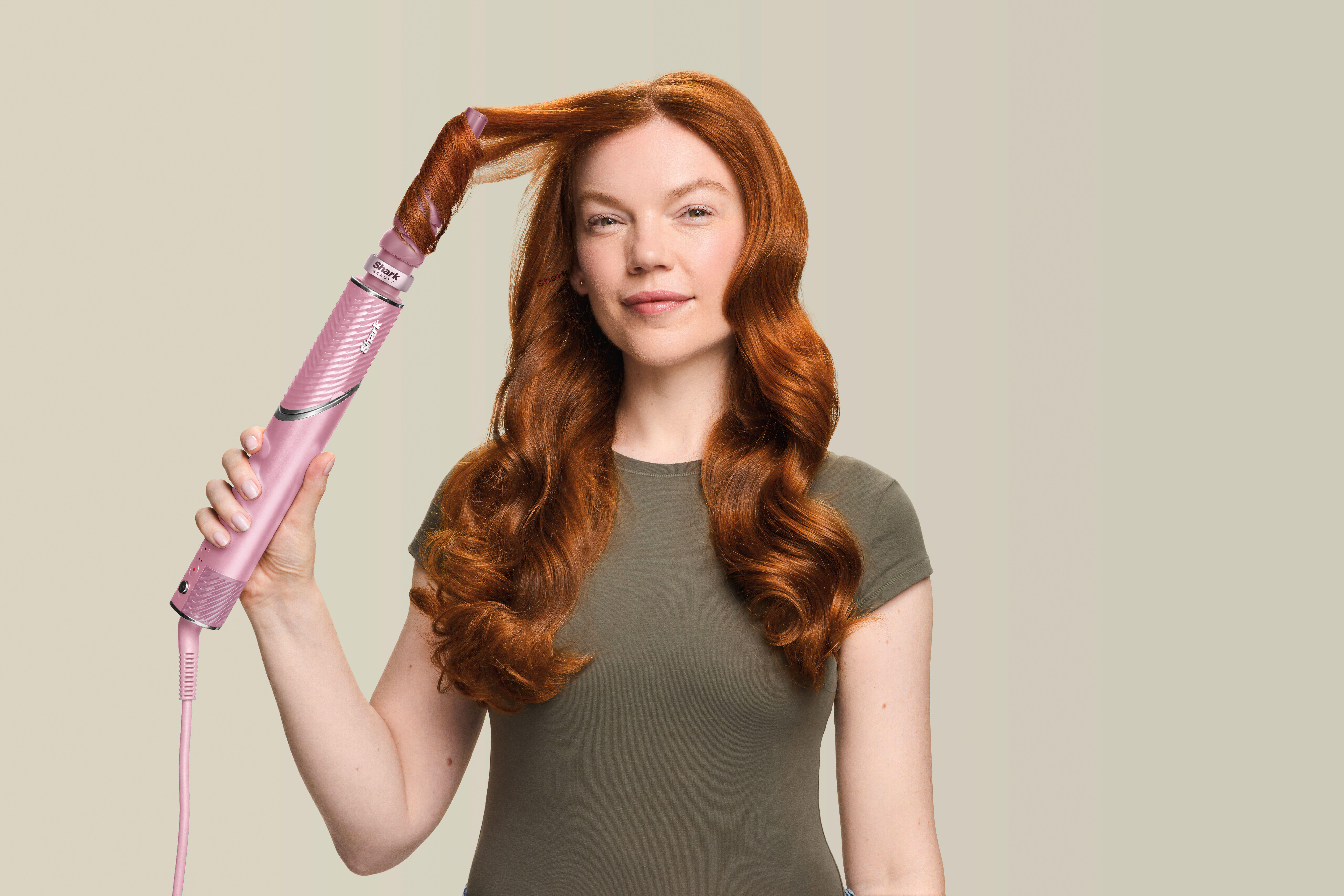 Eine rothaarige Frau hält einen rosafarbenen Lockenstab an eine Haarsträhne vor neutralem Hintergrund.