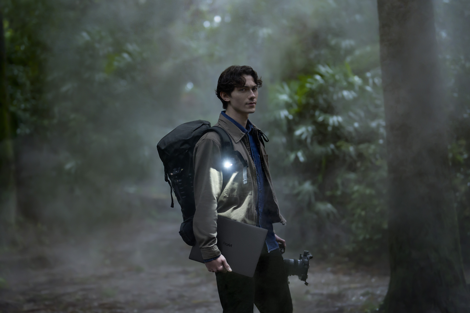 Un joven con mochila y portátil está en un bosque con niebla.