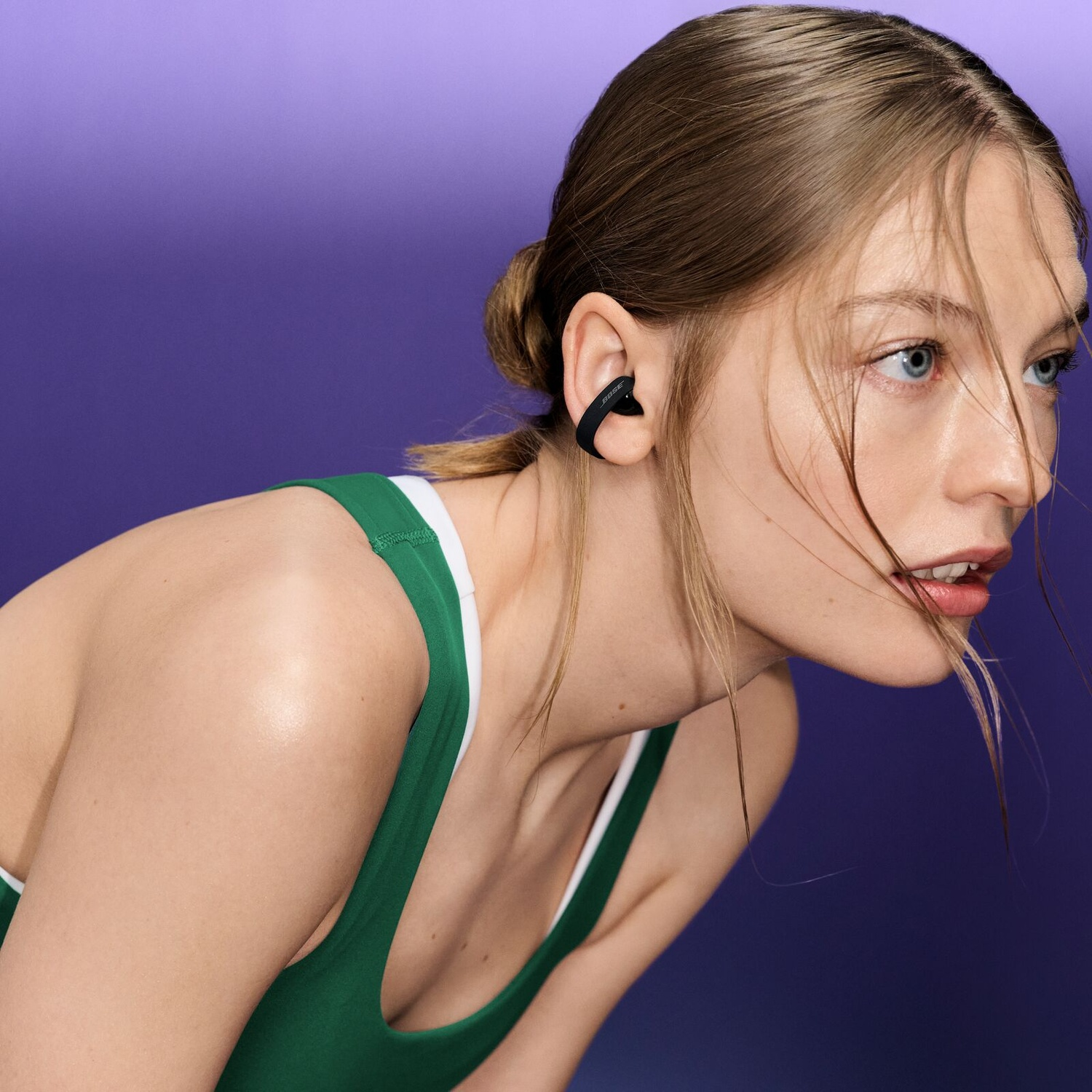 Une femme aux cheveux clairs avec un haut vert et un écouteur noir, sur fond violet uni.