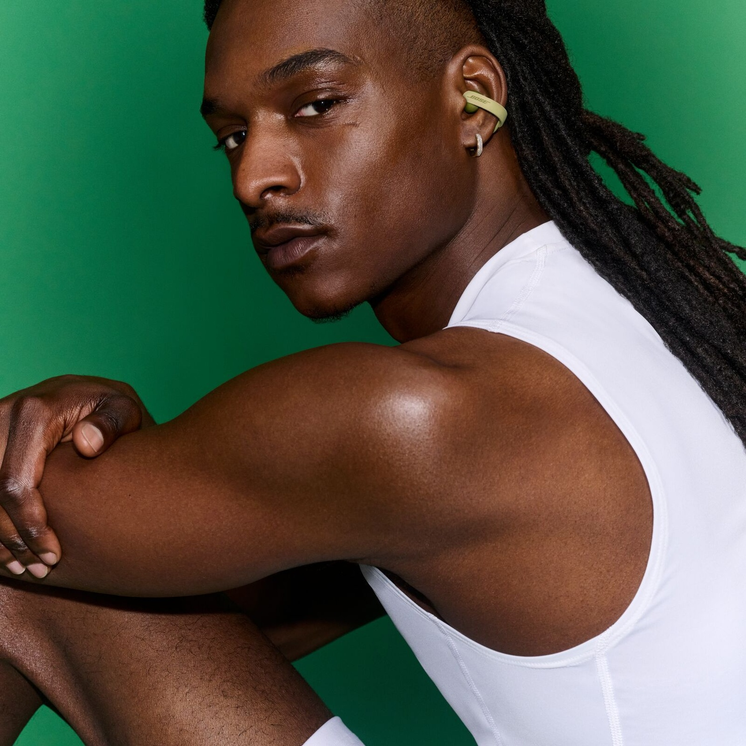 Un homme noir avec des dreadlocks et des boucles d'oreilles porte un débardeur blanc sur fond vert.