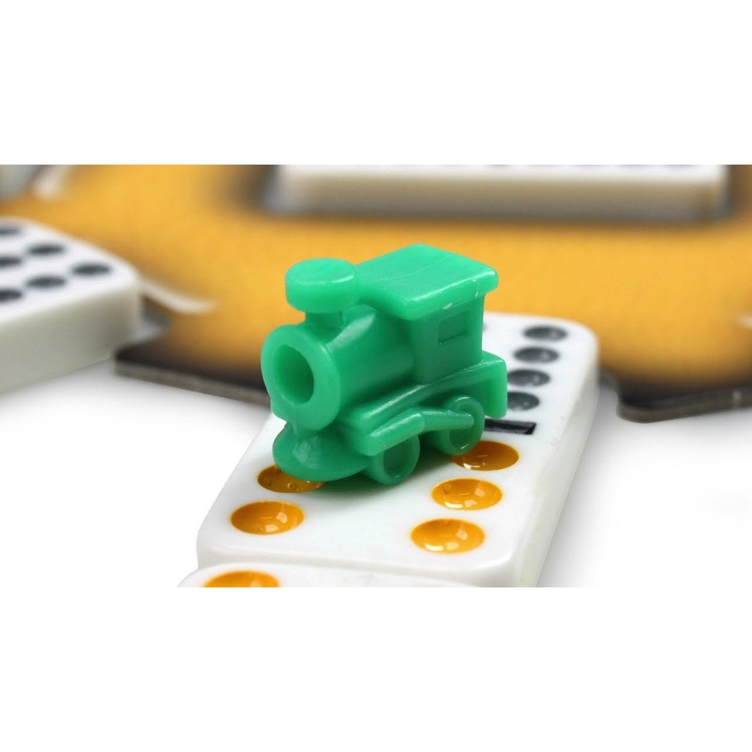 A small green plastic toy train sits on a white domino with yellow dots, against a blurred background.