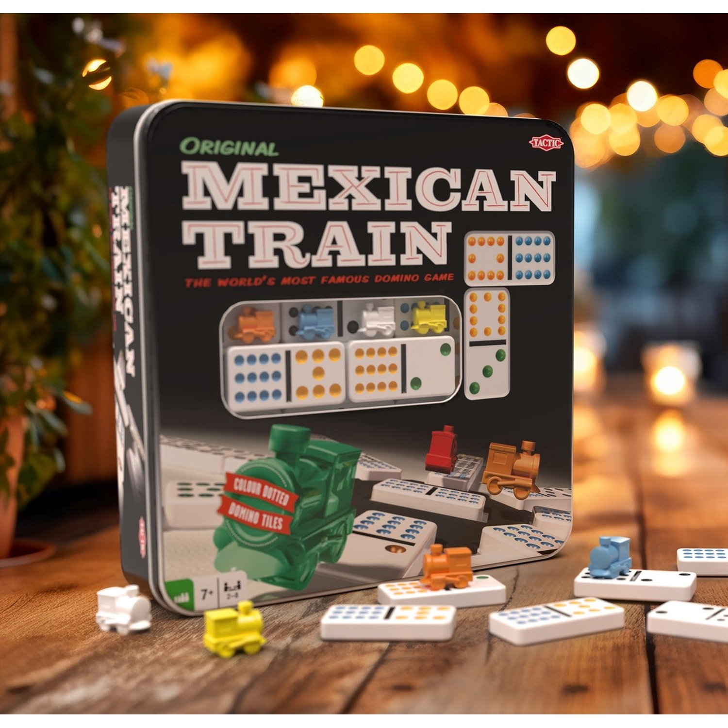 The box for Original Mexican Train dominoes sits on a wooden table, surrounded by domino tiles and small train pieces.
