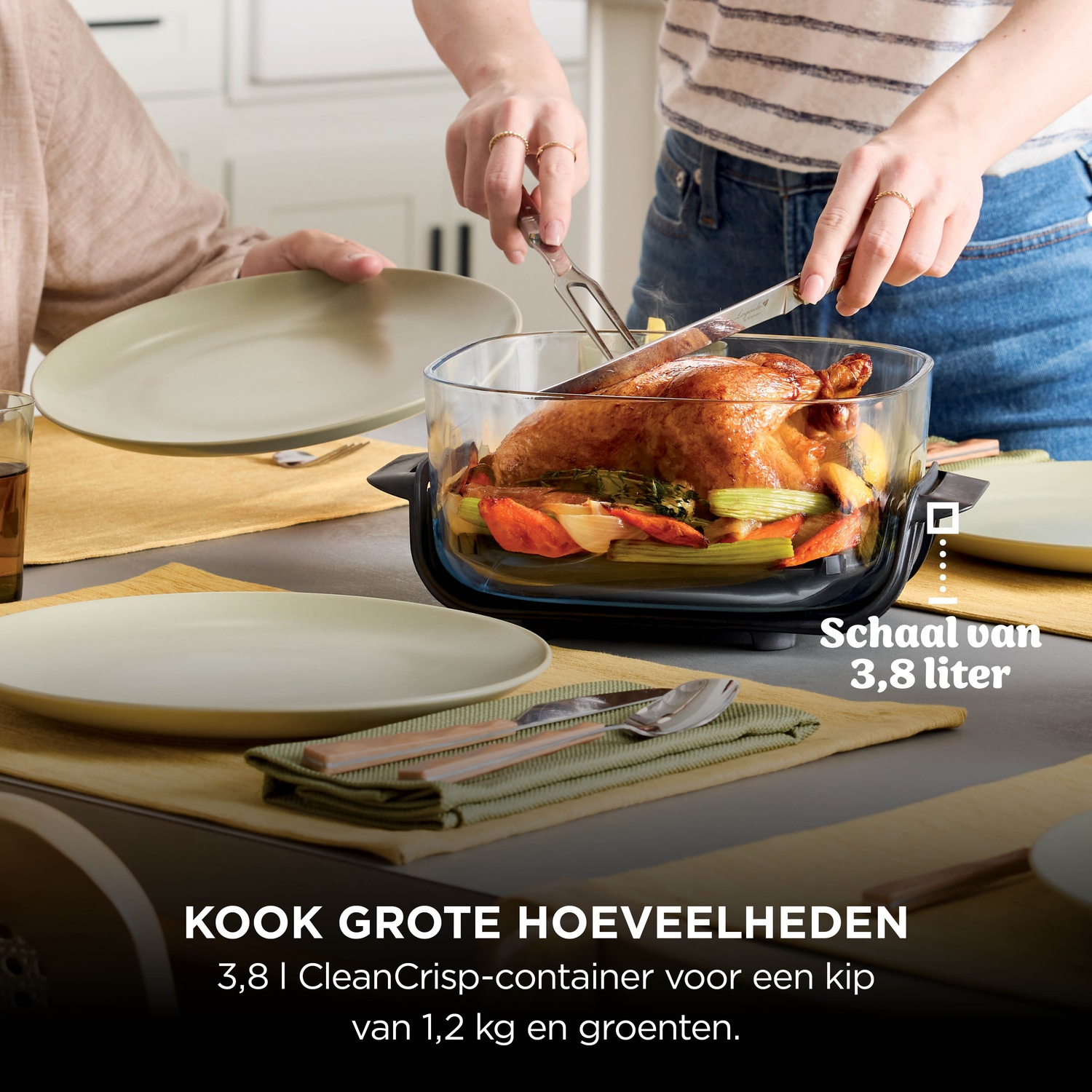 A person carves a whole roasted chicken with vegetables in a glass baking dish at a set table.