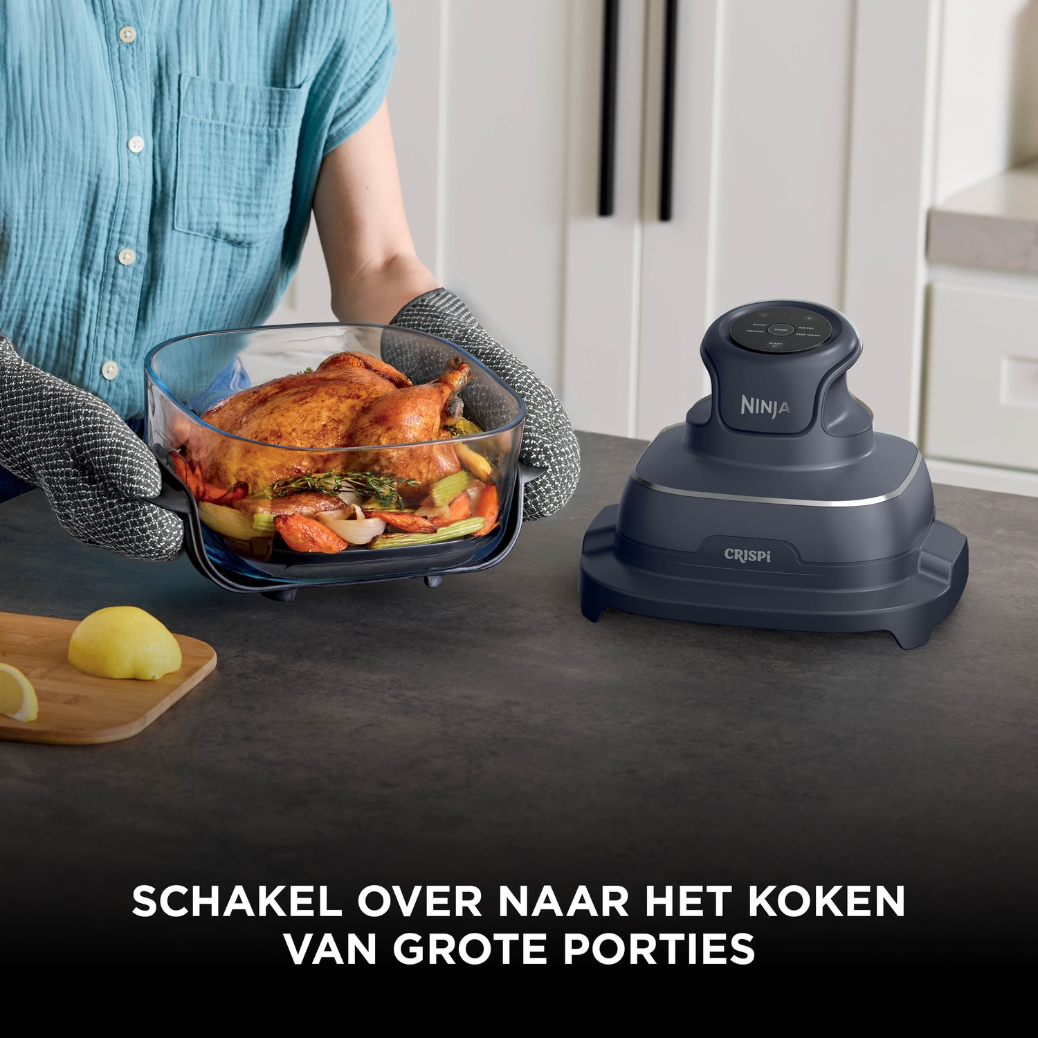 Person holding baked chicken in glass dish next to grey Ninja Crispi appliance on a kitchen counter.