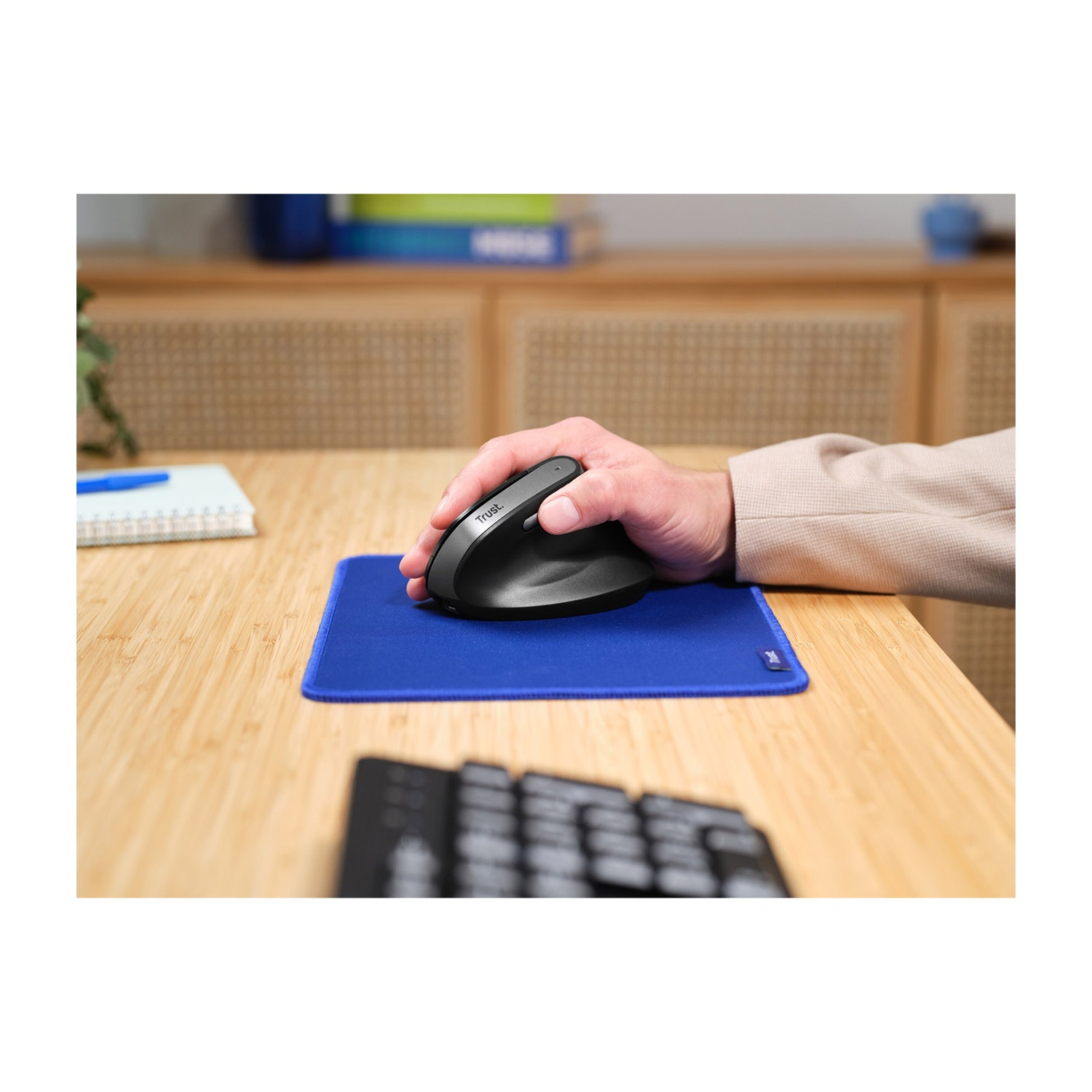 Una mano usa un mouse verticale nero su un tappetino blu su una scrivania di legno.