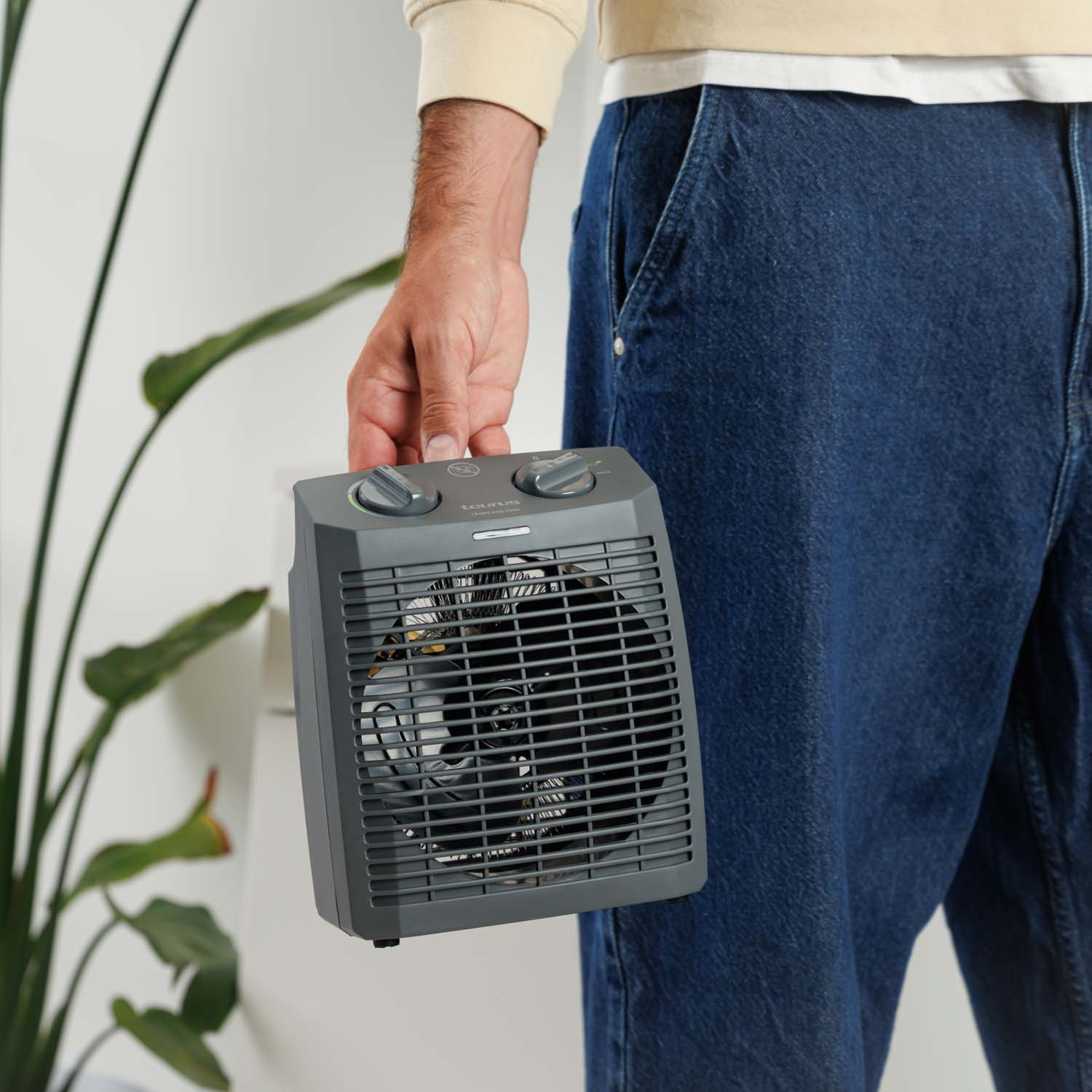Persona en jeans sostiene un calefactor portátil gris frente a una pared blanca y una planta.
