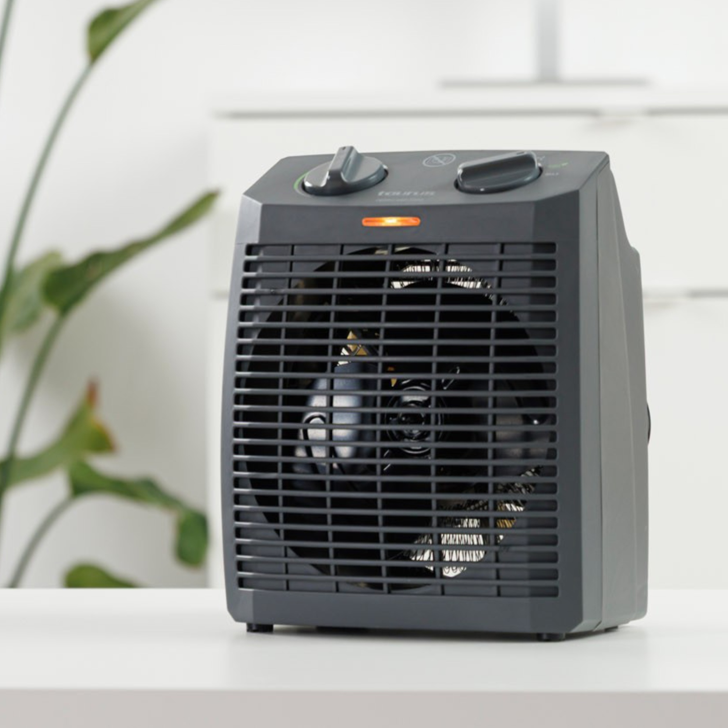 Un calefactor eléctrico gris sobre una superficie blanca, con dos mandos arriba y una luz de encendido naranja.