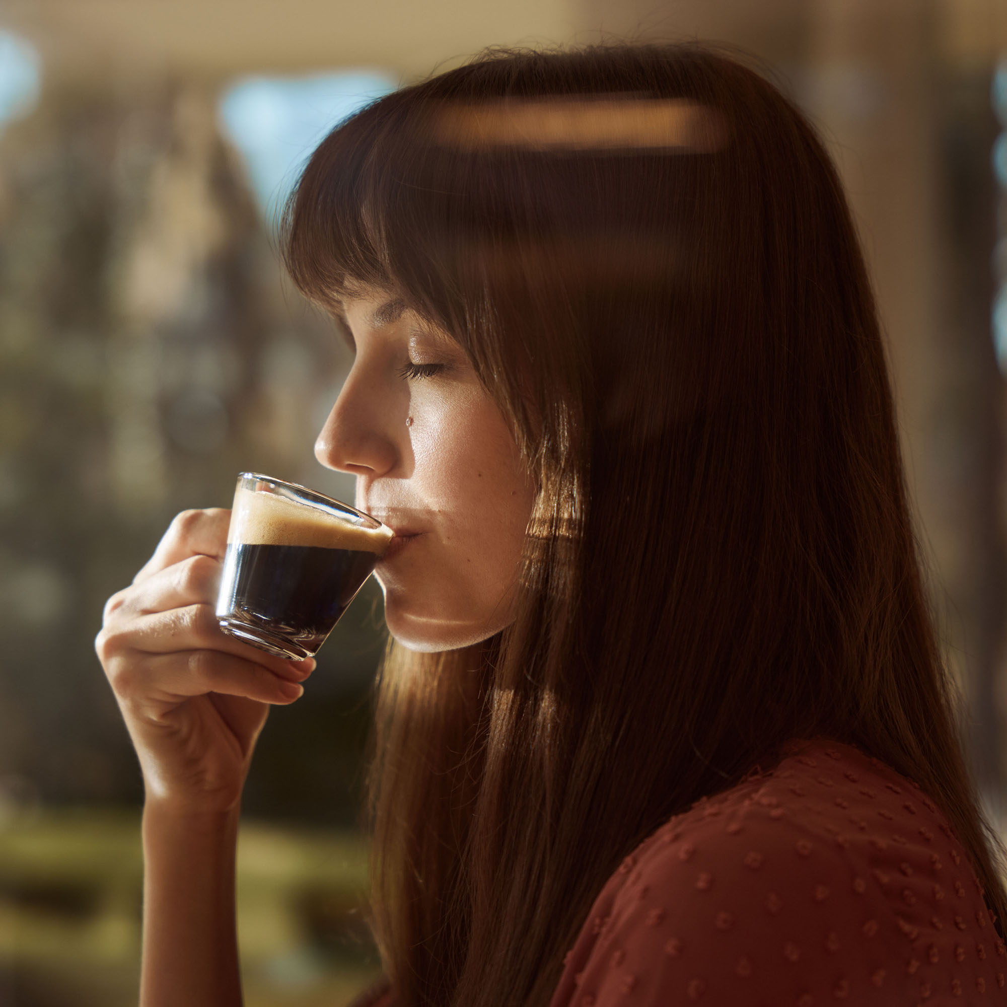Una donna bruna beve espresso da un bicchierino con calda luce interna.