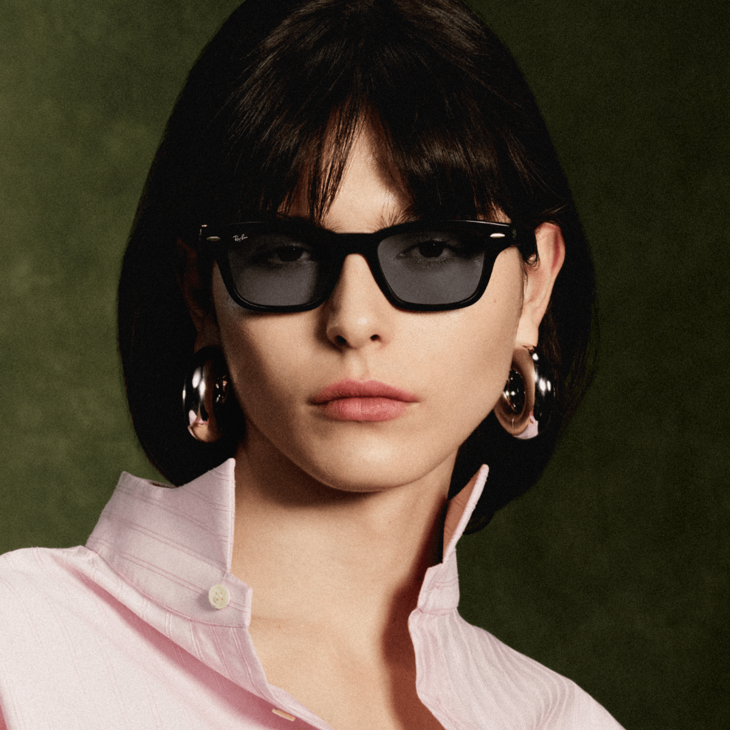 Una mujer de pelo castaño con gafas de sol negras, aros plateados y camisa rosa ante fondo oscuro.