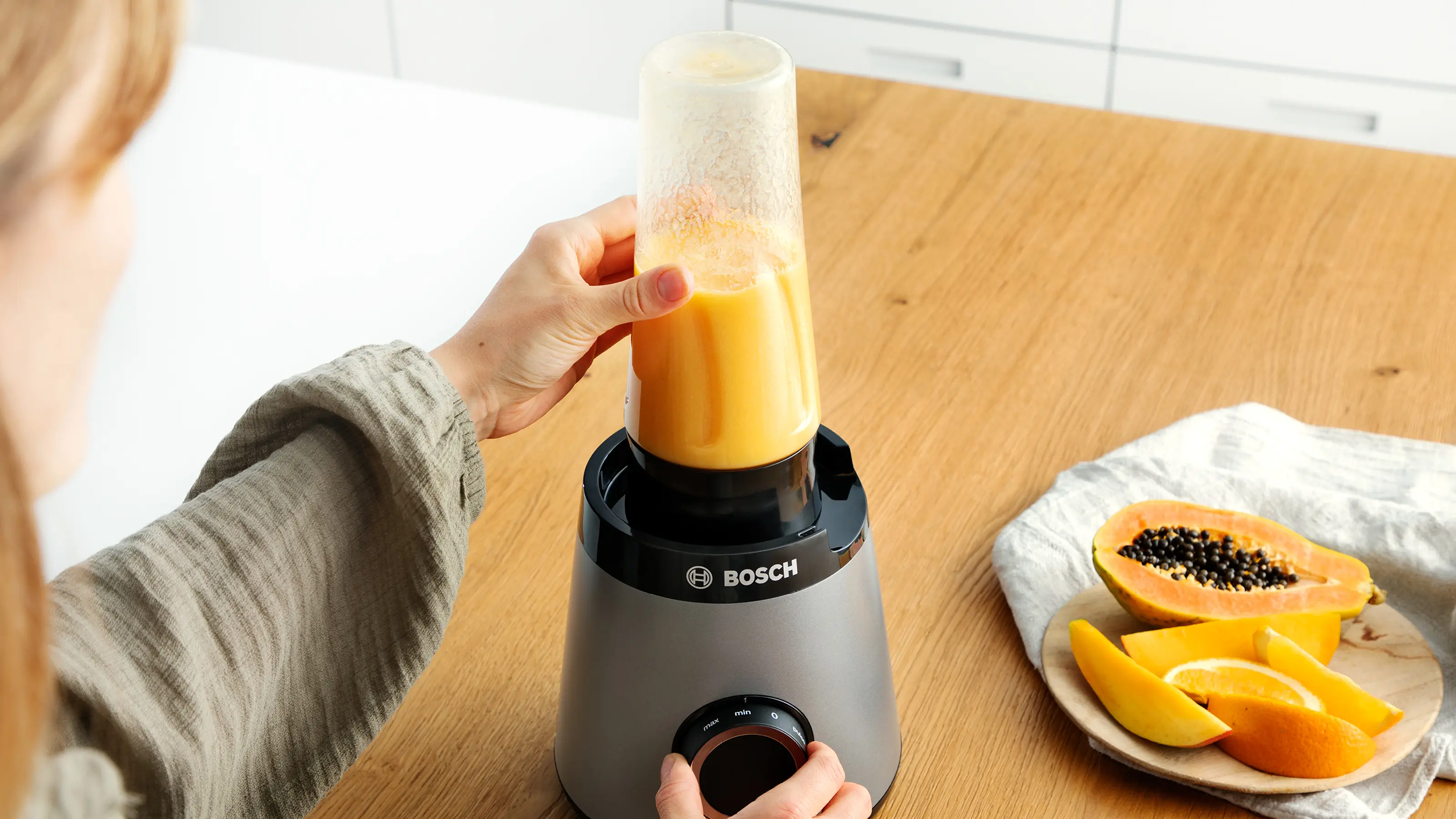 Egy személy egy szürke Bosch turmixgépet használ narancssárga smoothie-hoz, mellette friss gyümölcs.