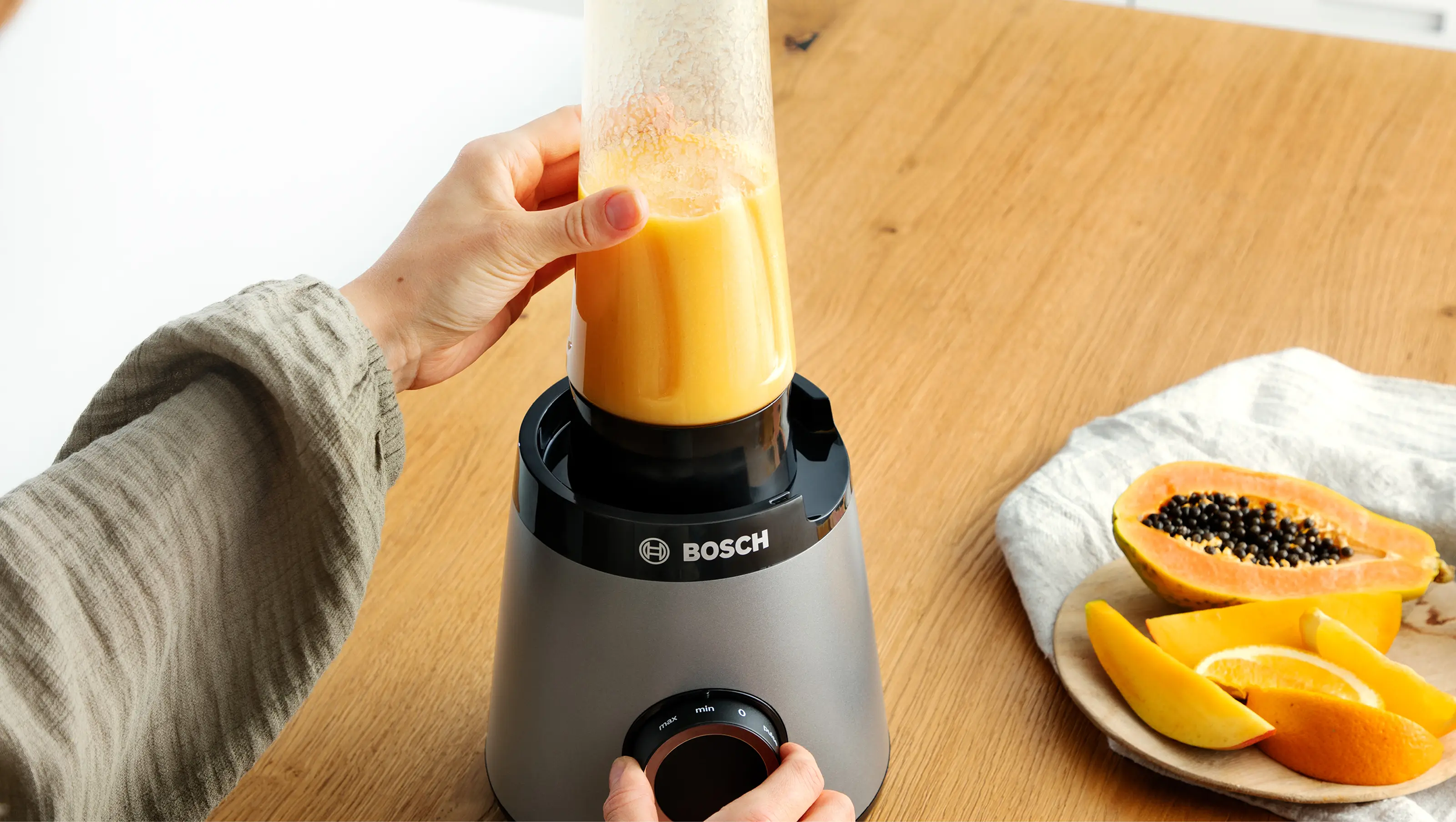 Egy személy narancssmoothie-s poharat tesz egy szürke Bosch turmixgépre egy faasztalon, szeletelt papaya és narancs mellett.