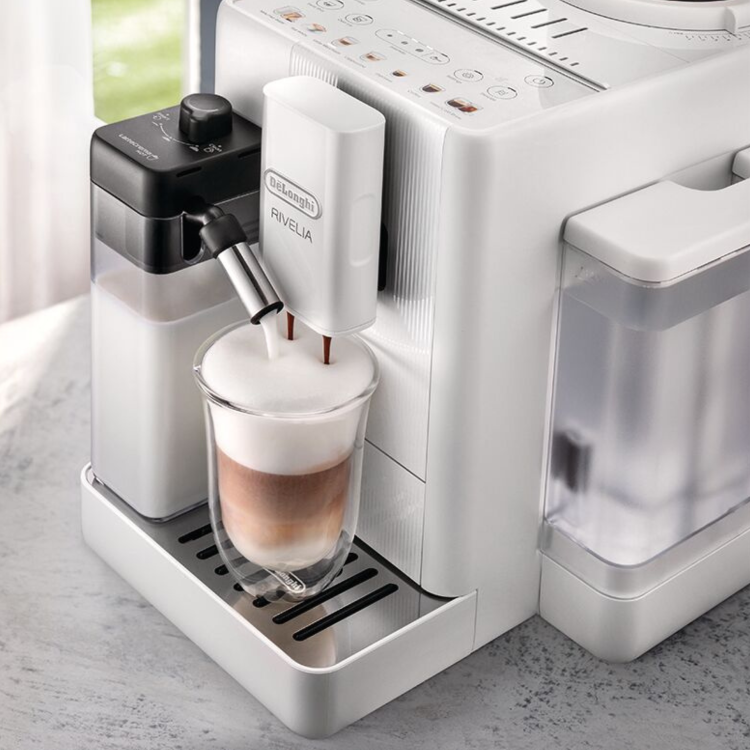 Una cafetera blanca DeLonghi Rivelia vierte leche y café en una taza de cristal.