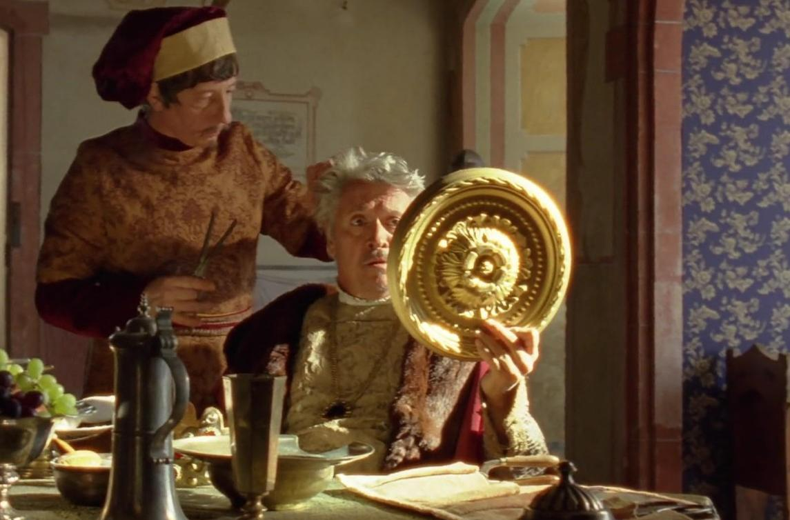 Ein Mann in mittelalterlicher Kleidung hält einen glänzenden goldenen Teller, während ein anderer mit Scheren hinter ihm steht.