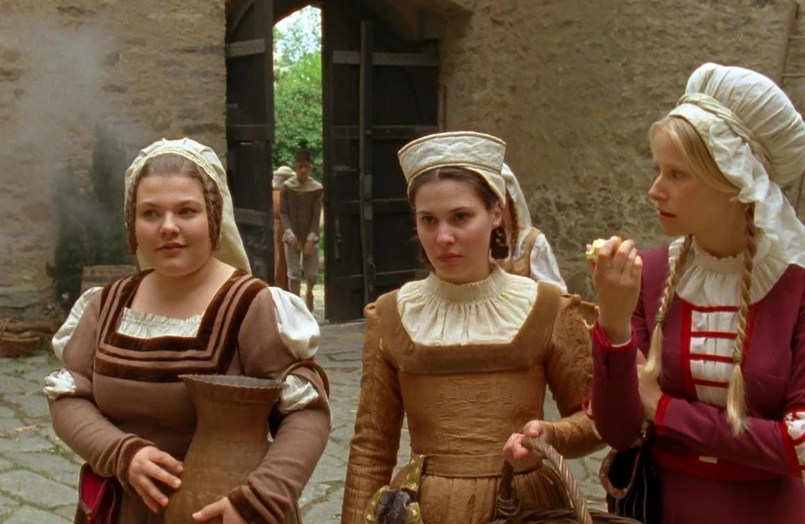 Drei Frauen in historischer Kleidung und Hauben stehen in einem Schlosshof, eine hält einen Krug, eine andere ein Körbchen.