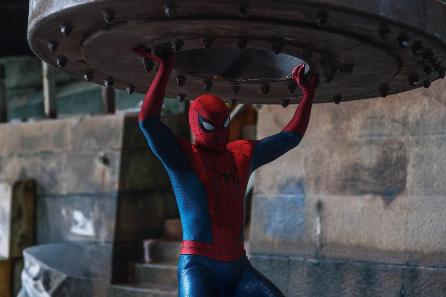 Spider-Man in rotem und blauem Kostüm hebt eine schwere, metallene Abdeckung in einem dunklen Raum an.