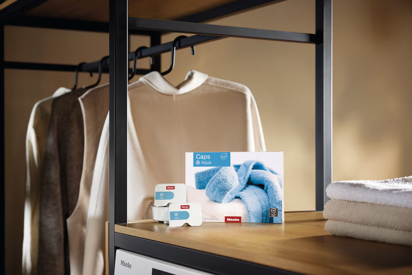 Miele Caps Aqua mosószer csomagolás fa polcon, háttérben ruhákkal.