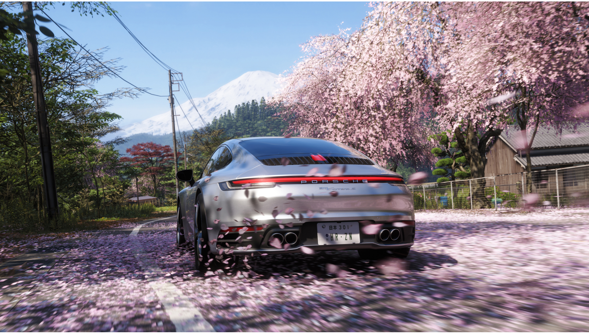 Silberner Porsche fährt auf einer Straße voller Kirschblüten, im Hintergrund blühende Bäume und ein schneebedeckter Berg.