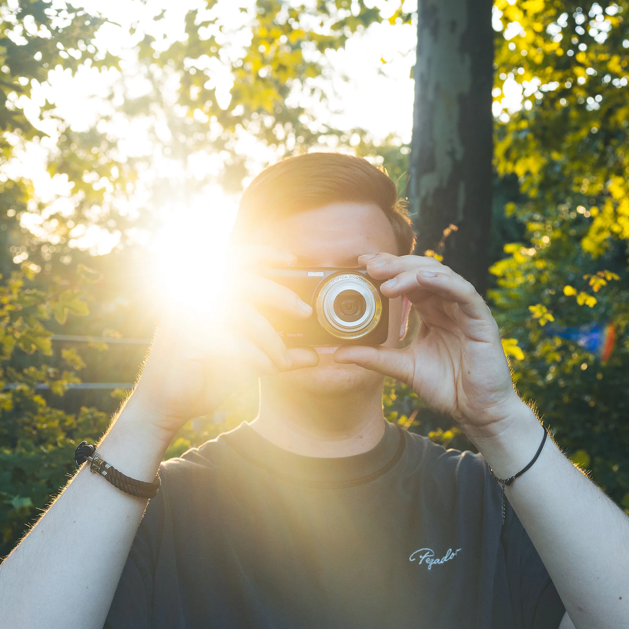 Ein Mann in einem T-Shirt hält eine Kamera vor sein Gesicht, während im Hintergrund Sonnenlicht durch Bäume scheint.