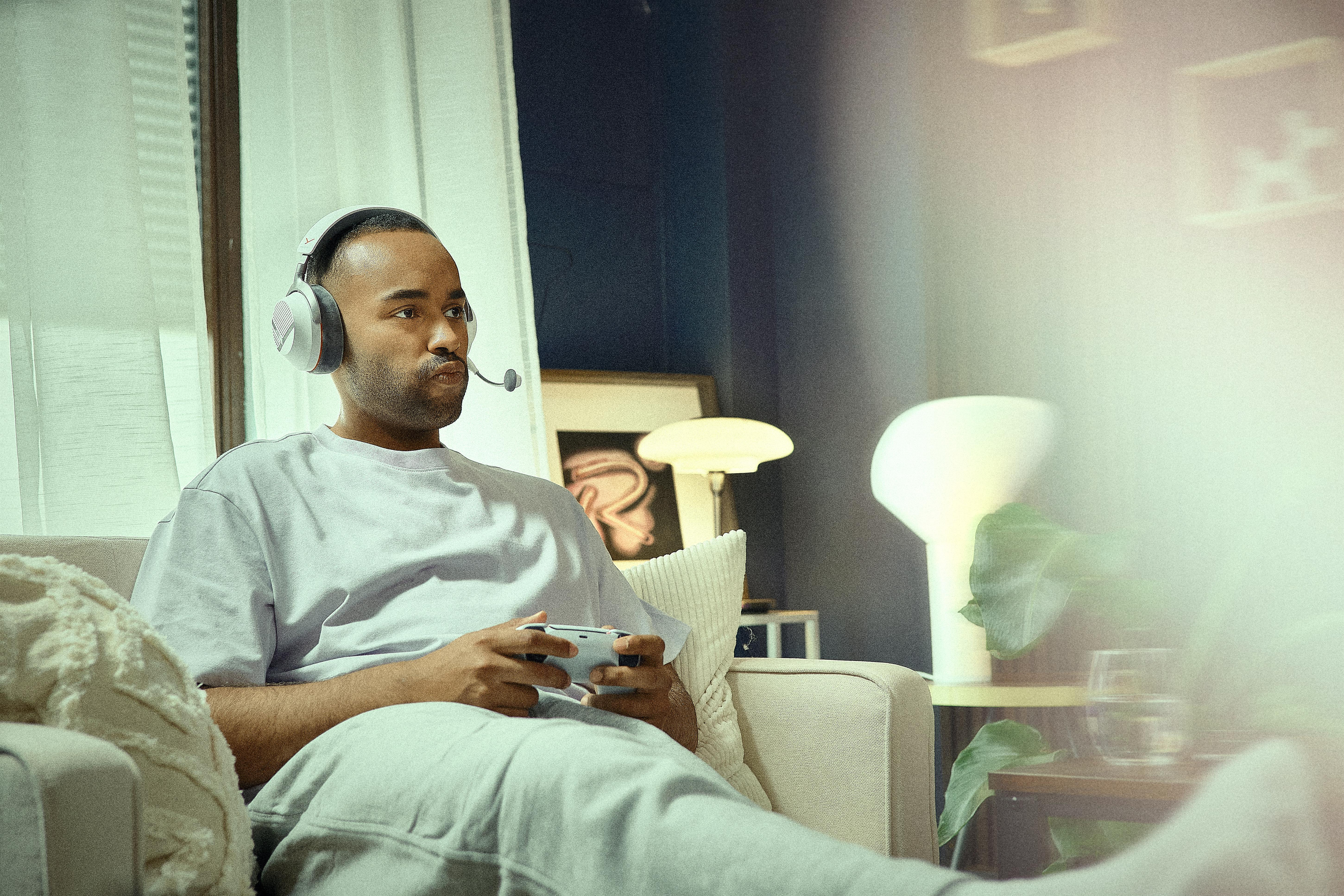 Ein Mann mit Kopfhörern und einem grauen Pullover sitzt auf einem Sofa und hält einen Controller in einem hellen Raum.