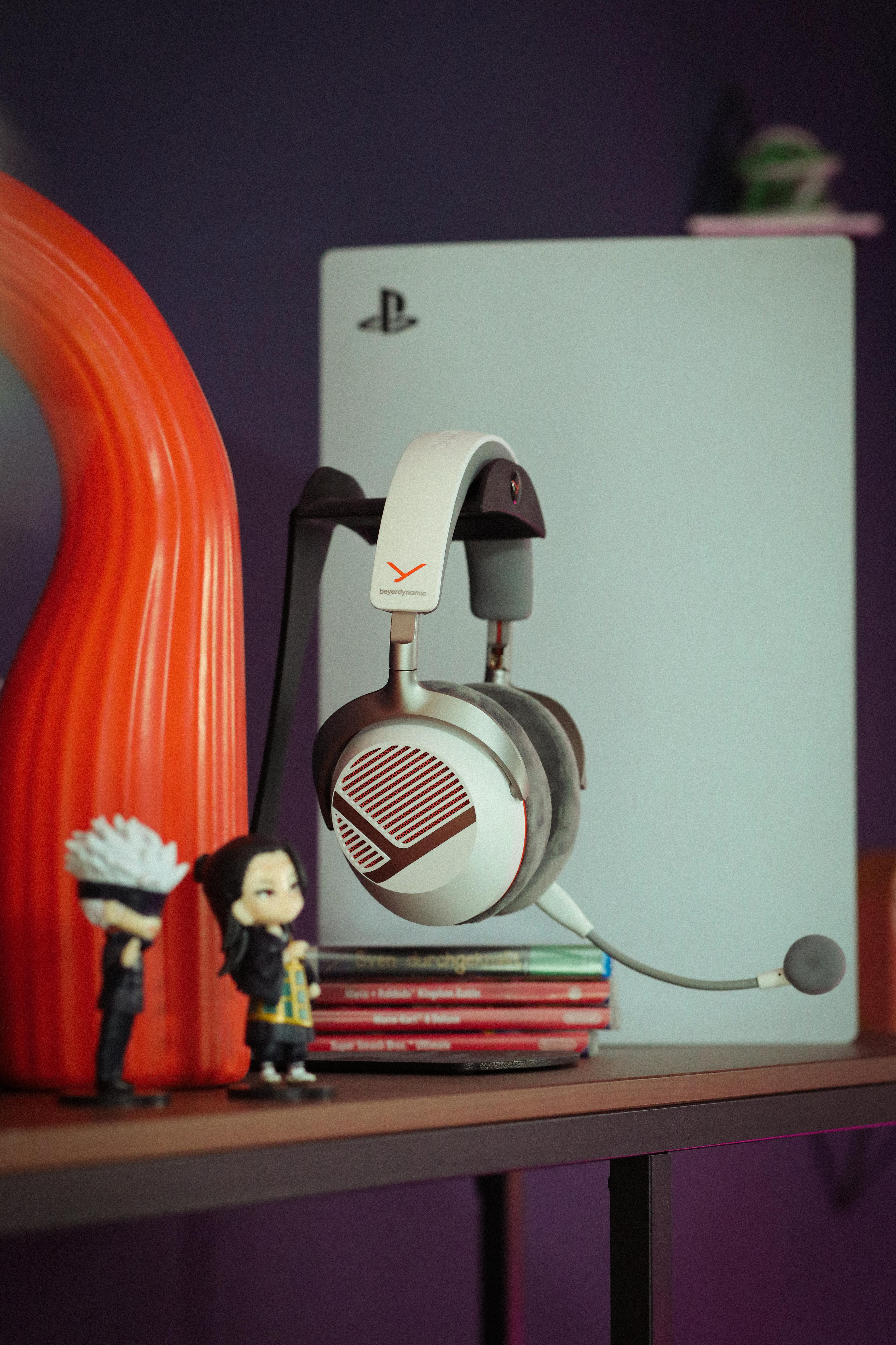 Weiße Kopfhörer auf einem Ständer vor einer PlayStation 5, Figuren und einer roten Skulptur auf einem Regal.