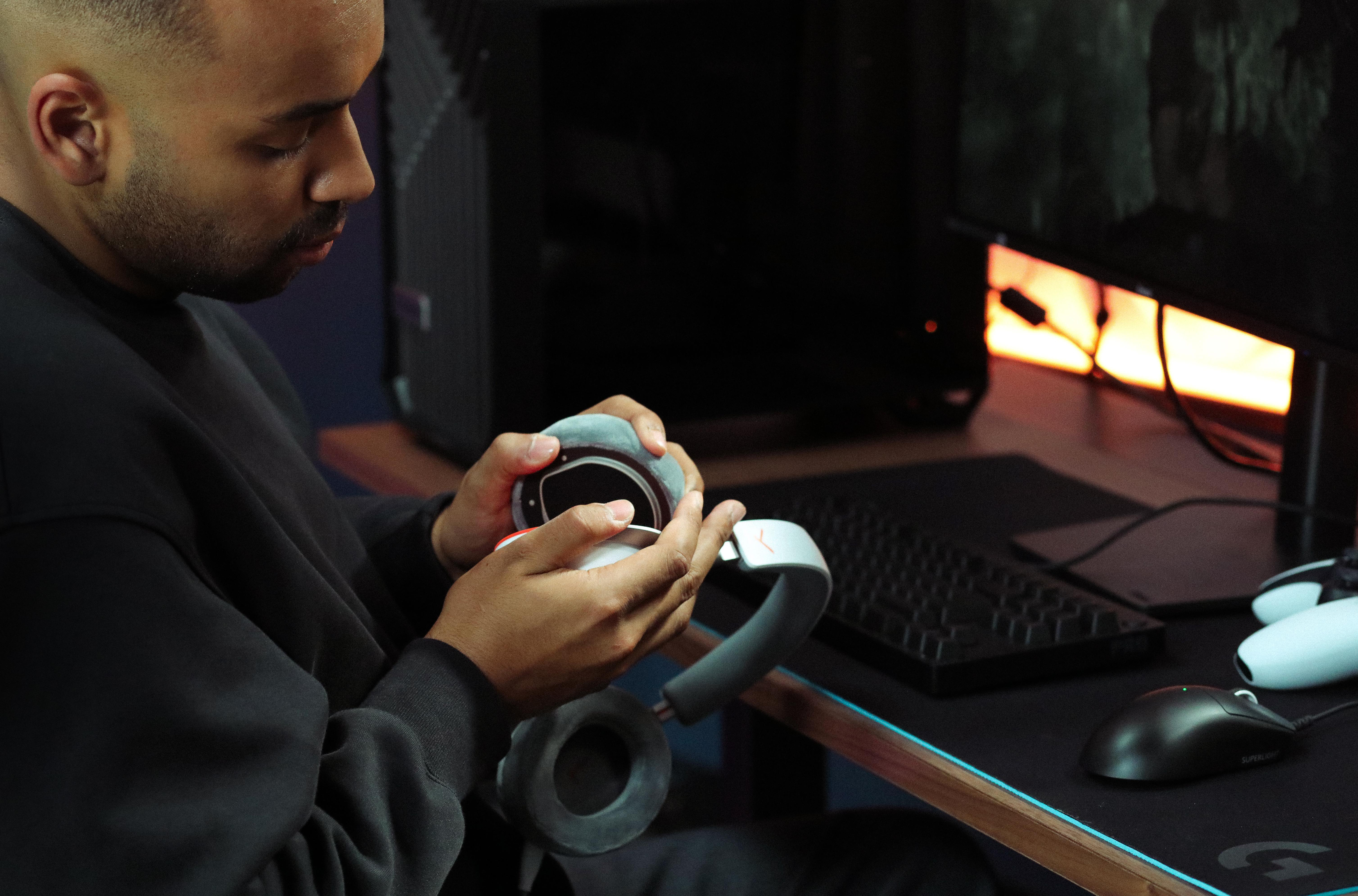Ein Mann im schwarzen Pulli repariert graue Kopfhörer an einem Schreibtisch mit Gaming-PC.