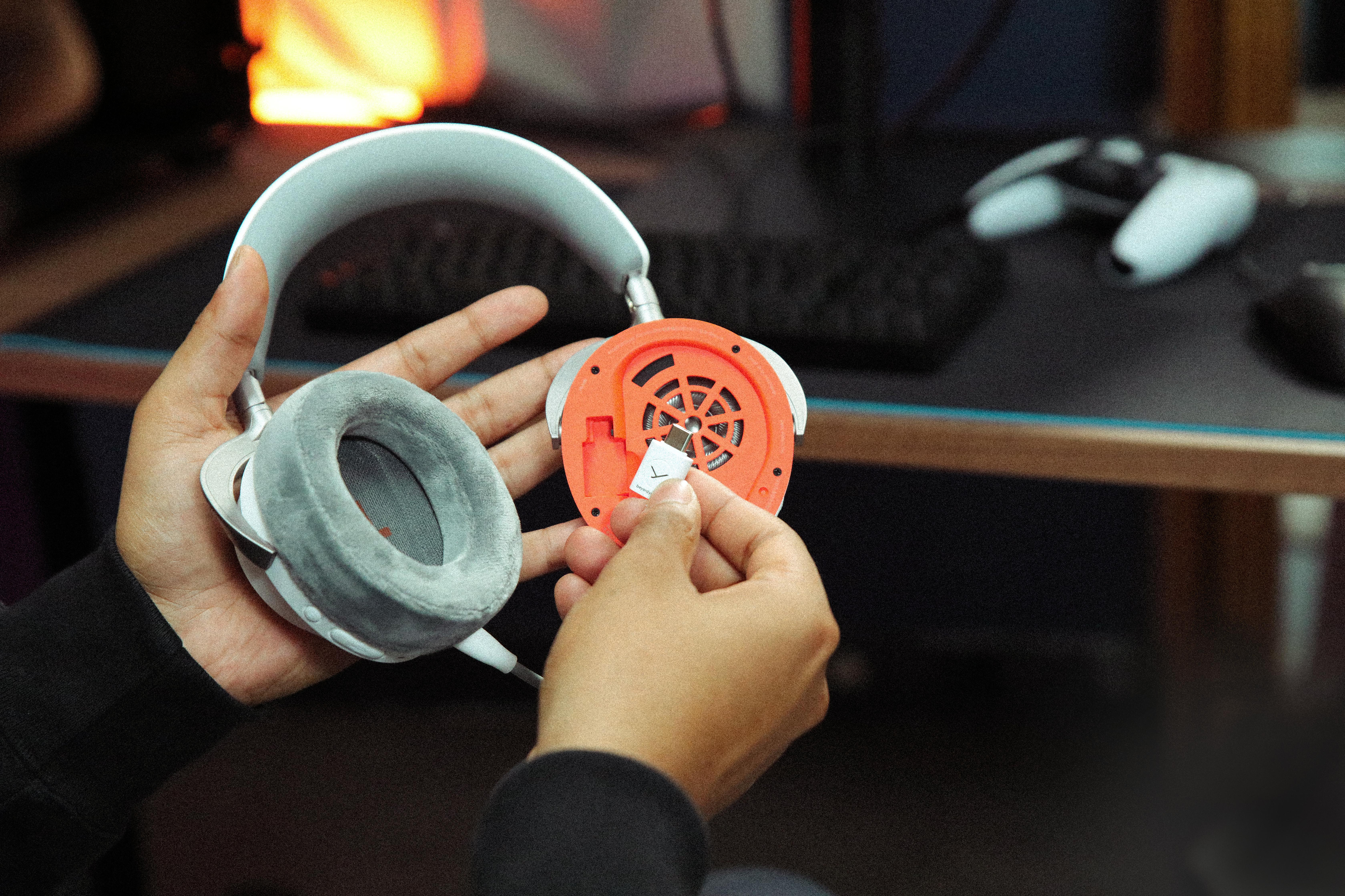 Hände halten weiß-orange Gaming-Kopfhörer mit einem USB-Dongle in der offenen orangen Ohrmuschel.