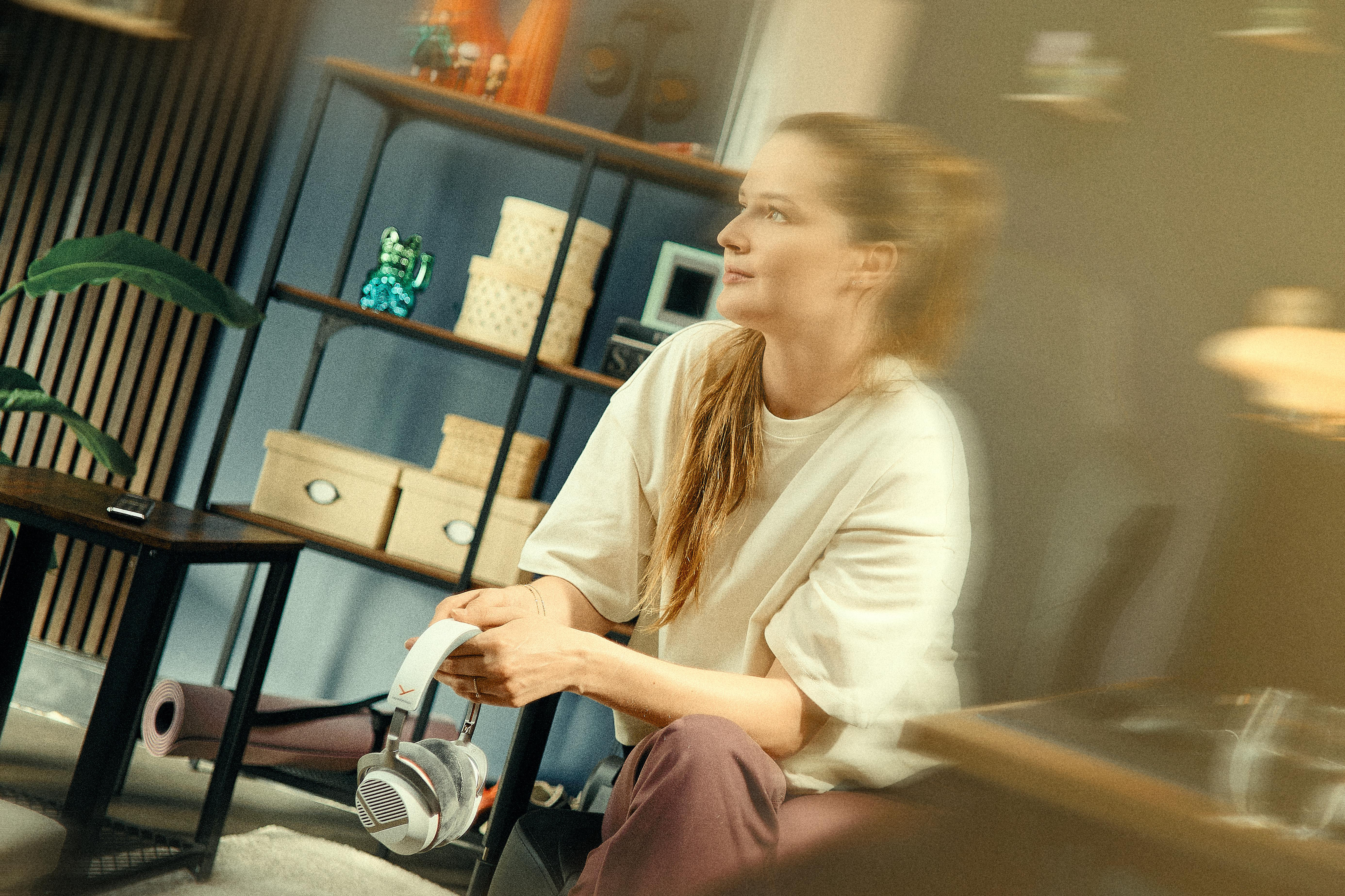 Eine Frau im weißen T-Shirt sitzt in einem Wohnzimmer und hält Kopfhörer in den Händen.