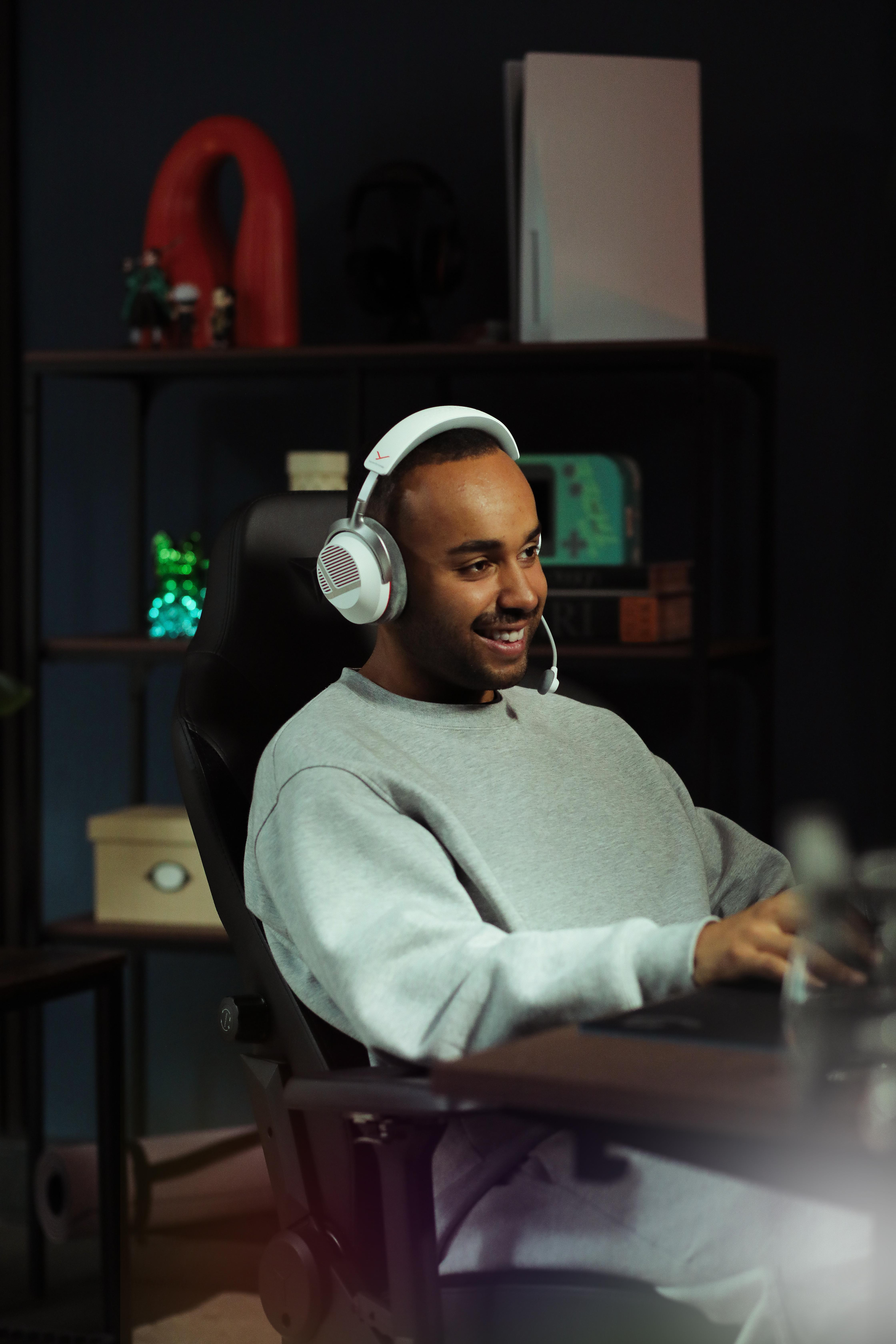 Ein lächelnder Mann mit weißen Kopfhörern sitzt in einem Gaming-Stuhl an einem Schreibtisch.