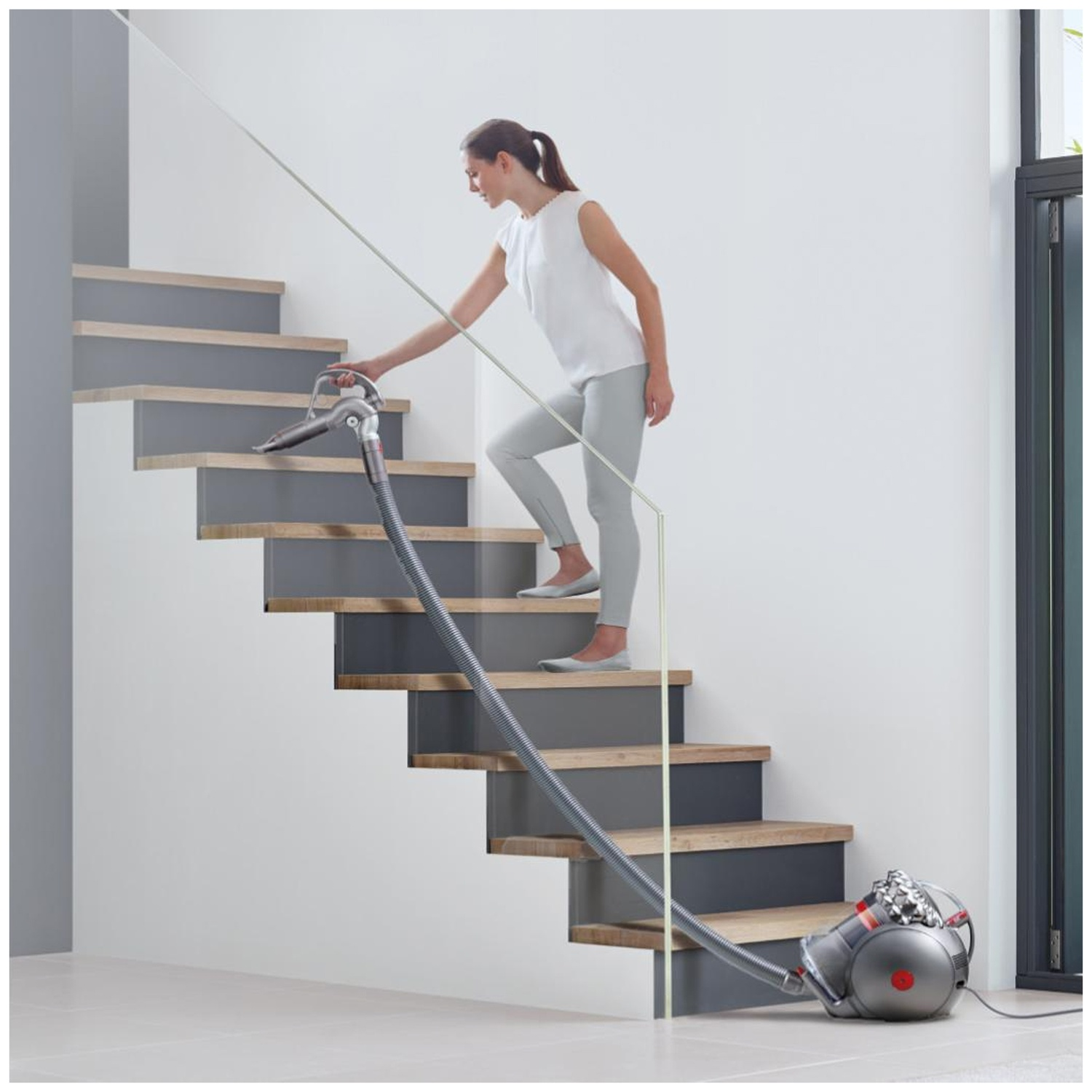 A woman in light clothing vacuums grey stairs with a canister vacuum cleaner.