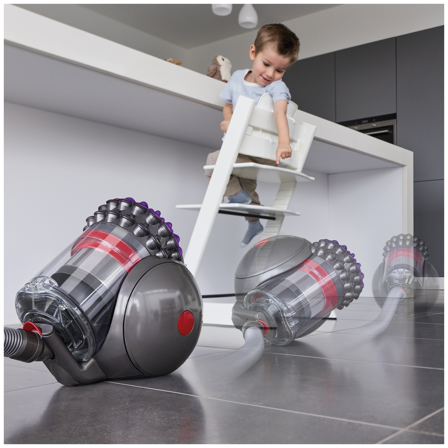 A grey Dyson canister vacuum cleaner moves across a tiled kitchen floor while a child watches from a high chair.