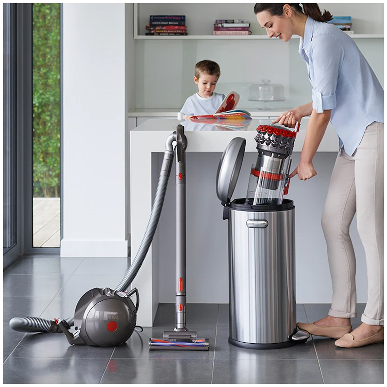 A woman empties a vacuum cleaner bin into a silver trash can in a kitchen while a boy reads.