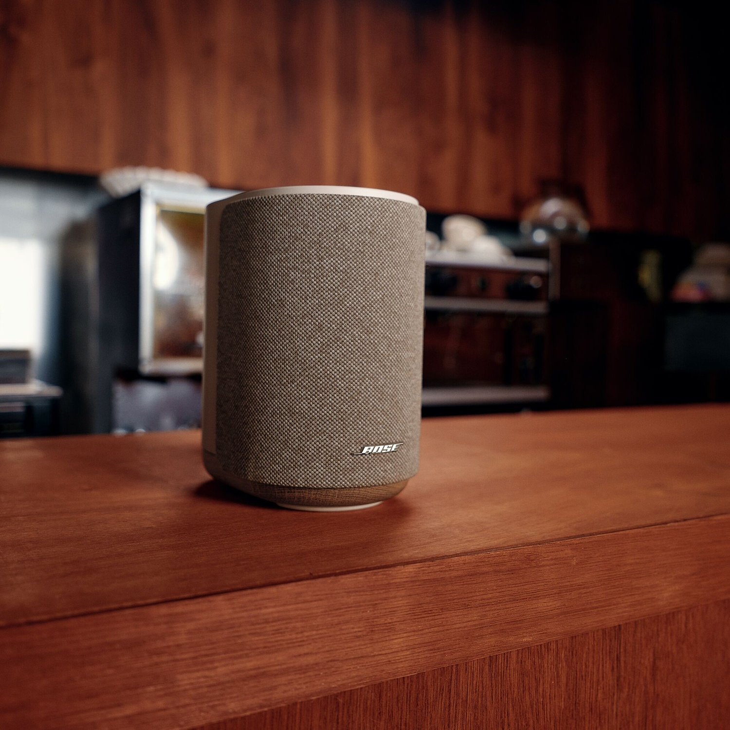A grey Bose speaker sits on a wooden counter in a kitchen with wood-paneled walls.