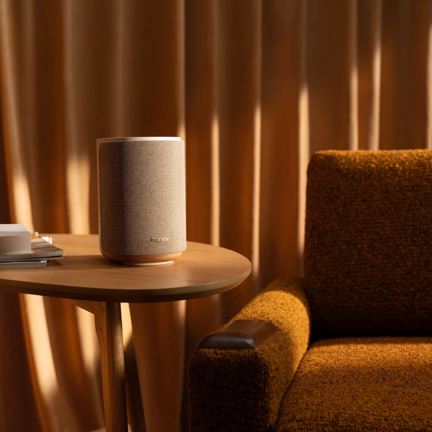 A cylindrical speaker on a wooden table beside a brown armchair against curtains.