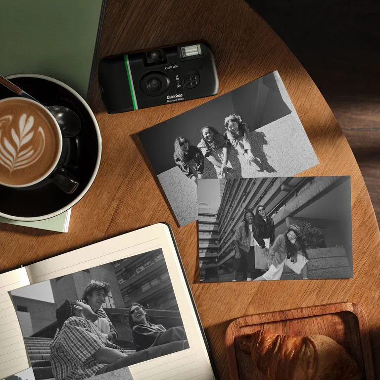 Table en bois avec café, appareil photo jetable, carnet ouvert, croissant et photos en noir et blanc.