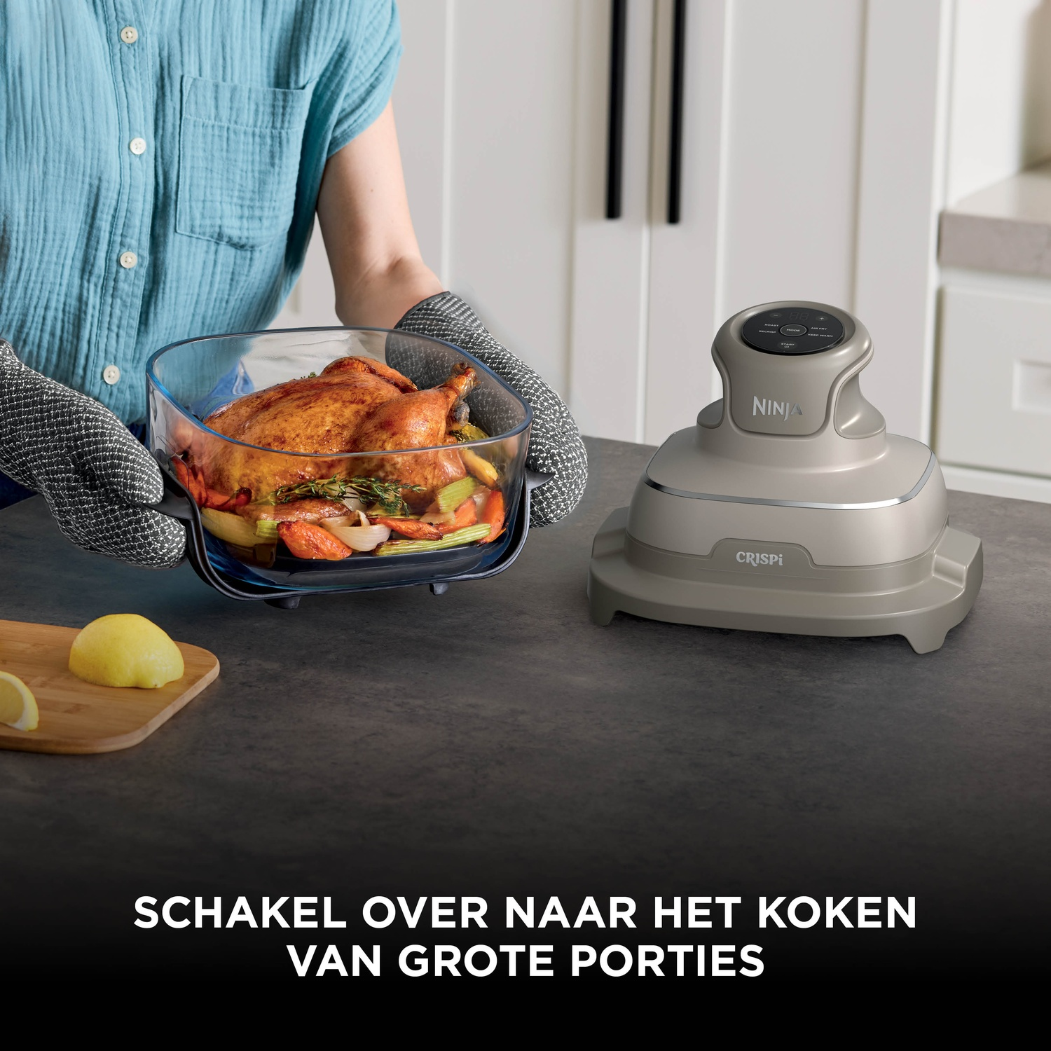 A person wearing oven mitts holds a glass dish with roast chicken and vegetables next to a Ninja cooker on a counter.