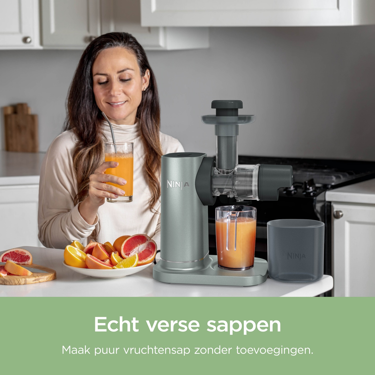A woman drinks fresh juice next to a gray Ninja juicer on a kitchen counter with citrus fruits.