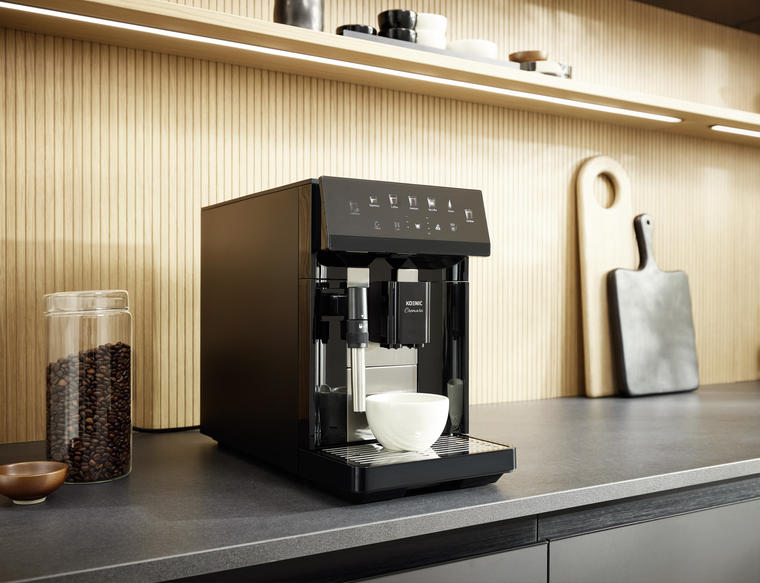 Eine schwarze Kaffeemaschine steht auf einer Küchentheke mit Kaffeebohnen und Schneidebrettern im Hintergrund.