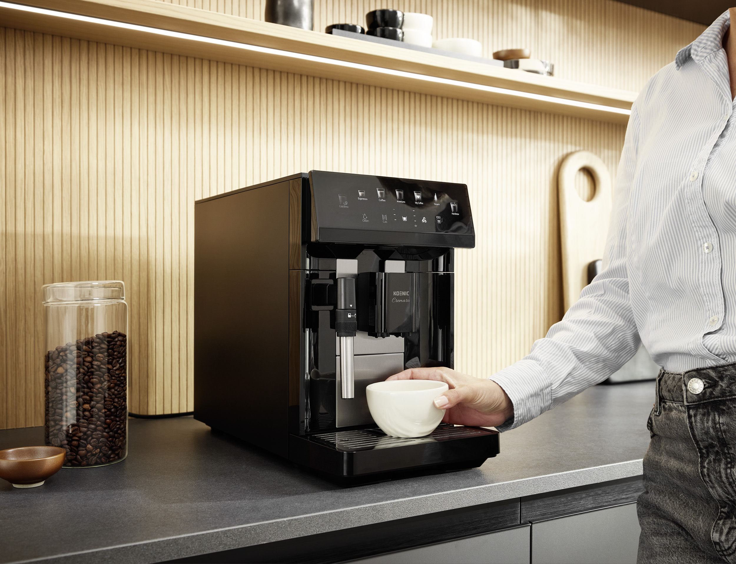 Eine Person stellt eine weiße Tasse vor einen schwarzen Kaffeevollautomaten in einer modernen Küche.